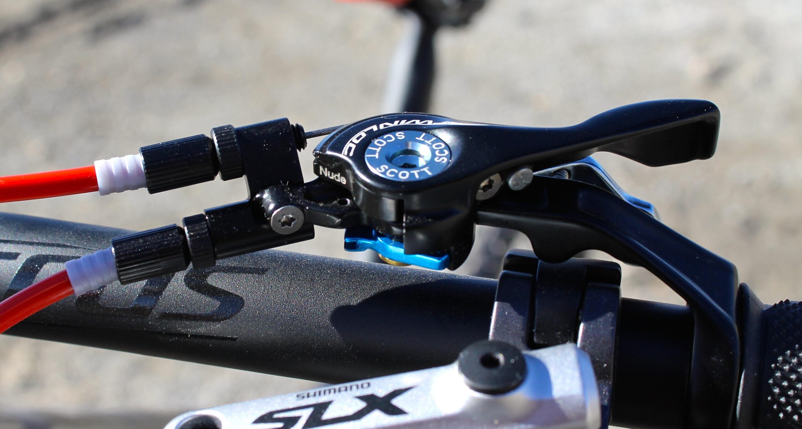 Scott Genius 930: Close-up image of a bicycle handlebar featuring a hydraulic brake lever with red and white cables connected. The lever is mounted on a sleek black handlebar, with part of a silver brake component visible below. The background is softly blurred, emphasizing the details of the brake lever and cables.