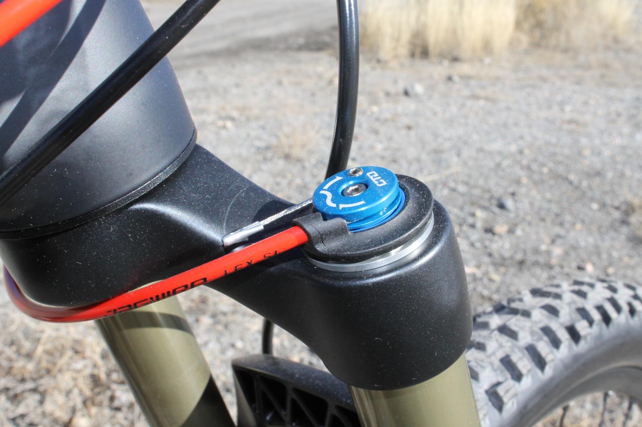 Scott Genius 930: Close-up view of a mountain bike fork's top tube, featuring a blue adjustment knob and a red cable. The background shows a gravel surface and sparse vegetation, indicating an outdoor setting.