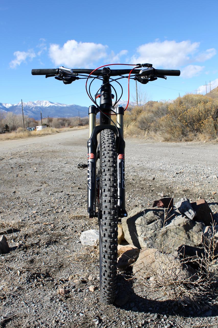 Scott Genius 930: A mountain bike viewed from the front, standing on a gravel path with a background of mountains and a clear blue sky. The bike features thick, knobby tires, suspension forks, and handlebars, with a few scattered rocks nearby.
