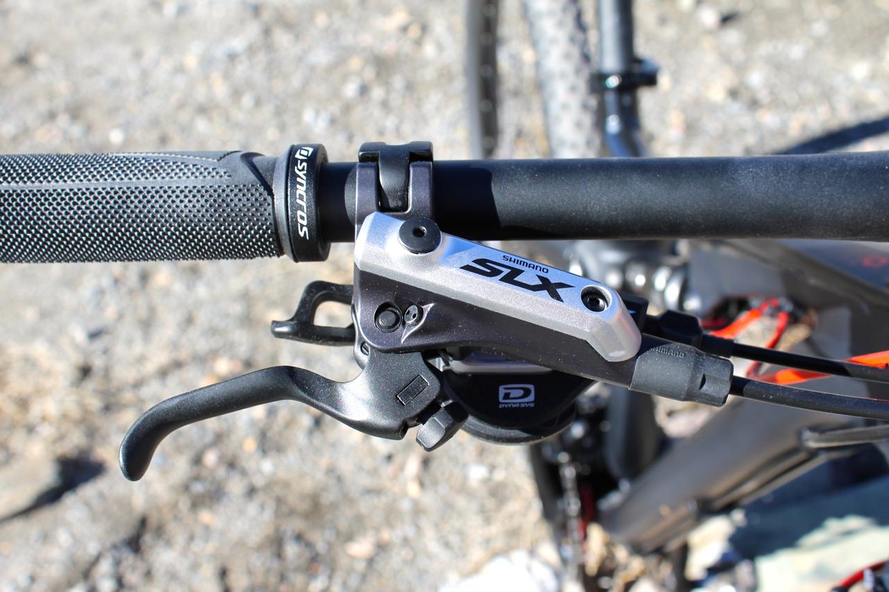 Scott Genius 930: Close-up of a bicycle handlebar featuring a Shimano SLX gear shifter, black rubber grip, and brake lever. The background shows a gravel surface.