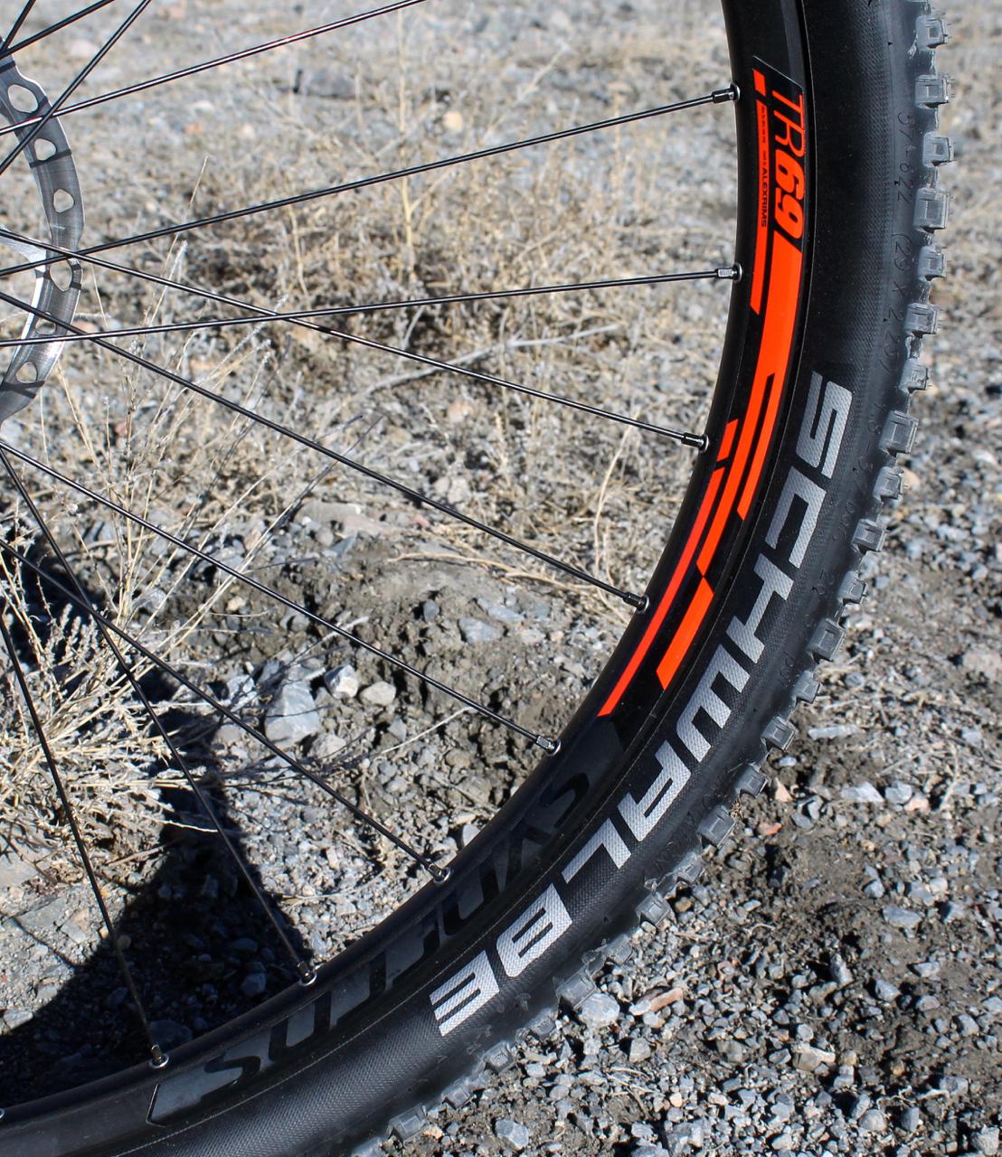 Scott Genius 930: A close-up view of a mountain bike wheel, featuring a black tire with a prominent logo in white reading "SCHWALBE" and an orange accent. The rim is partially visible, showcasing spokes and a disc brake system against a rugged, gravelly background with sparse vegetation.