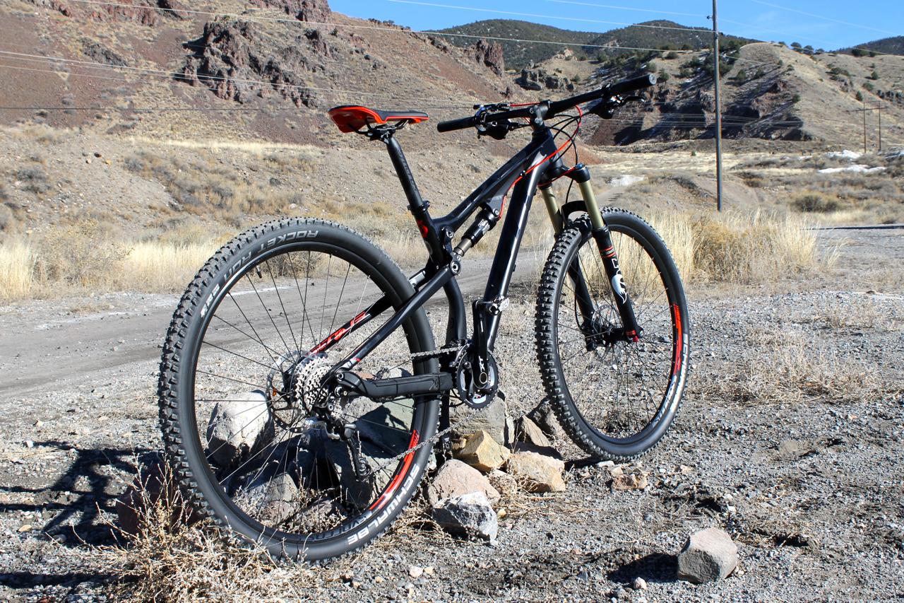 Scott Genius 930: A black and red mountain bike is positioned on a gravel path with rocky terrain and sparse vegetation. In the background, hills and a clear blue sky are visible, indicating an outdoor setting ideal for biking. The bike features thick tires, a suspension system, and a sleek design.