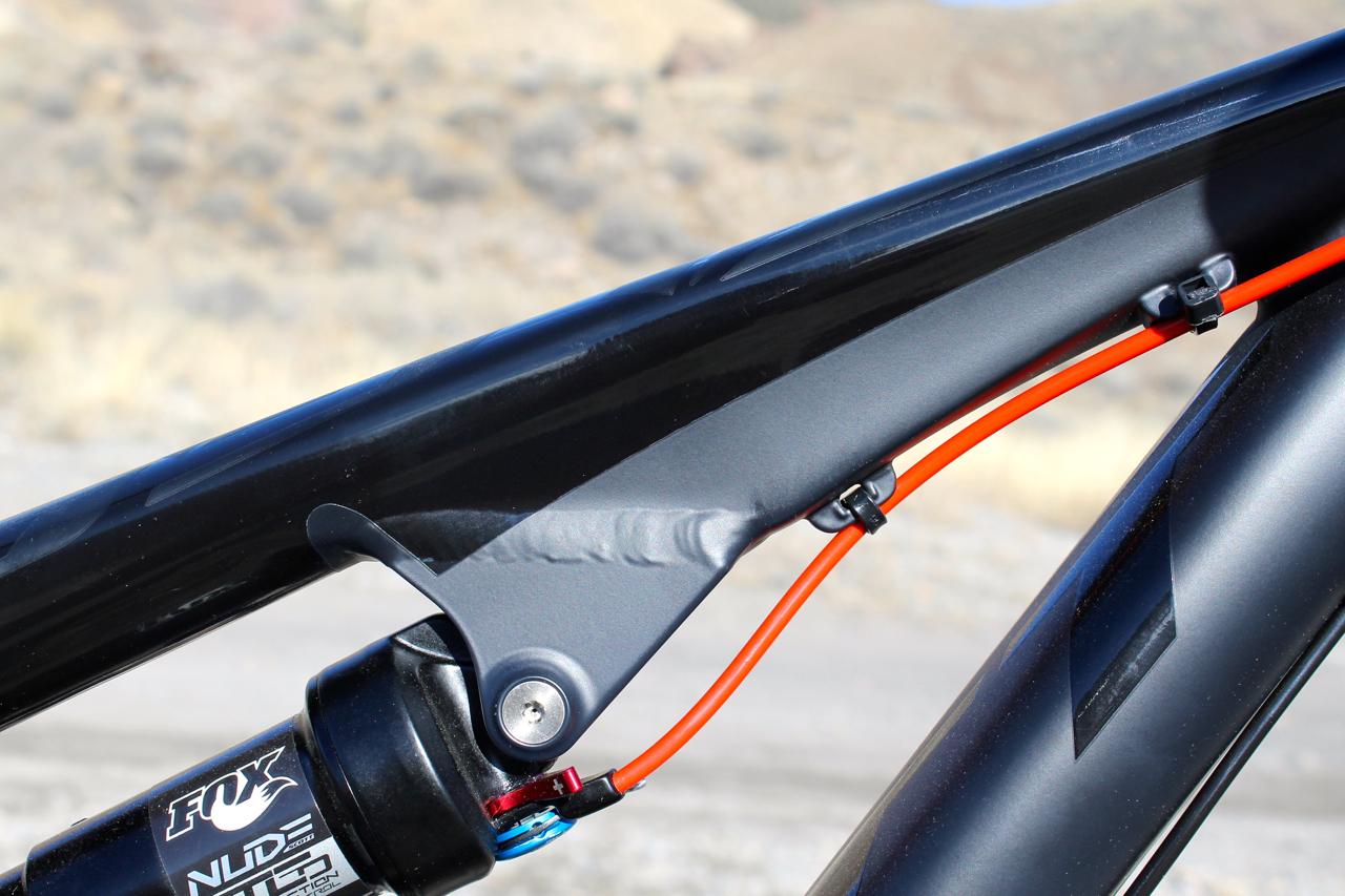 Scott Genius 930: Close-up of a bicycle frame detail featuring a gray suspension link and an orange cable. The frame shows a sleek design with a matte finish, while the background is blurred, suggesting an outdoor setting.