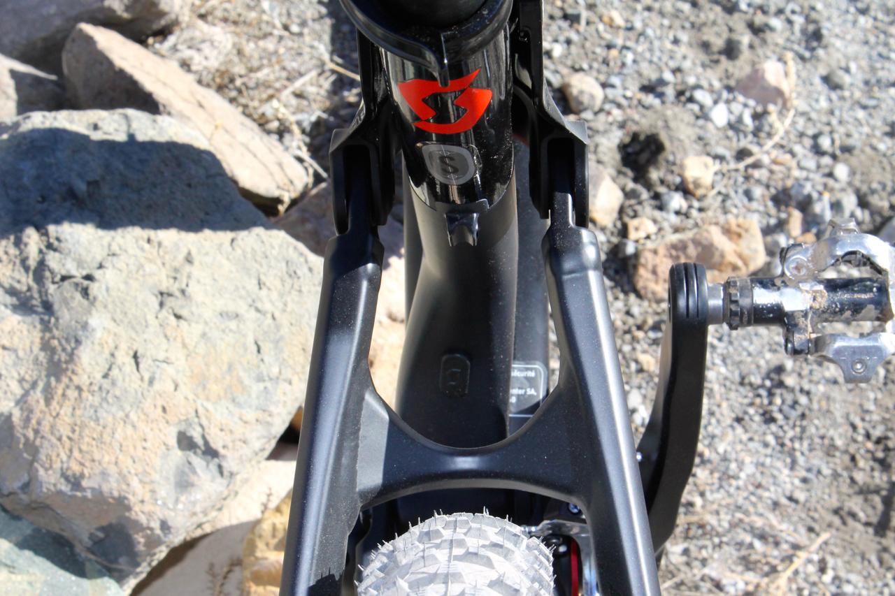Scott Genius 930: Close-up view of a black mountain bike frame showcasing the bike's bottom bracket area, with a distinct red logo on the seat tube. The bicycle's pedal is visible on the right, and the terrain features rocky ground.