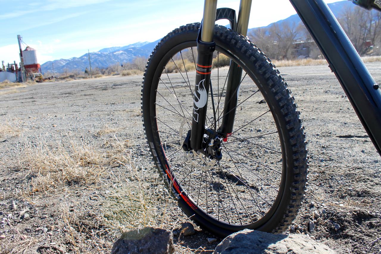 Scott Genius 930: Close-up view of a mountain bike front wheel and suspension fork, resting on a gravel surface with dry grass and rocks. In the background, a clear blue sky and distant mountain range can be seen.