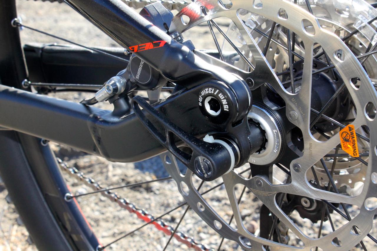 Scott Genius 930: Close-up view of a bicycle's rear wheel assembly, featuring the derailleur, chain, and disc brake components. The image captures the intricate machinery and chain setup, highlighting the Shimano branding and the metallic disc brake. The background is outdoors, with a gravel surface visible.