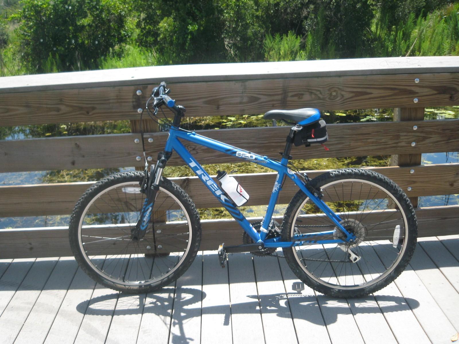 Trek 820: A blue mountain bike leaning against a wooden railing on a bridge, with a scenic view of greenery and water in the background. The bike has visible tires and a water bottle holder, suggesting it’s ready for a ride.