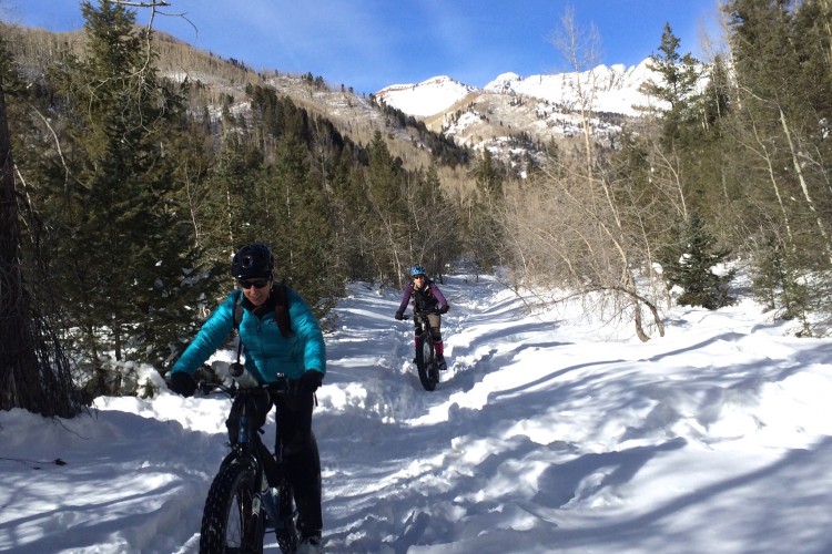 Two cyclists riding fat bikes on a snowy trail through a forested area, with snow-covered mountains visible in the background under a blue sky. One cyclist is wearing a turquoise jacket and the other is dressed in darker colors. The path is surrounded by trees, giving a sense of wilderness and adventure.