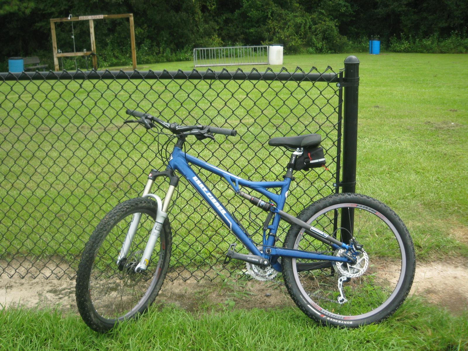 Gary Fisher HiFi Plus: A blue mountain bike leaning against a black chain-link fence, with grassy fields and playground equipment visible in the background.