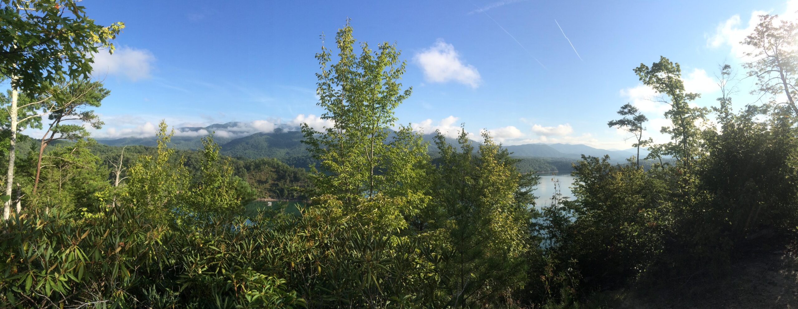 A panoramic view of lush green mountains under a blue sky with fluffy clouds, framed by vibrant trees in the foreground. The landscape features a serene body of water reflecting the surrounding scenery. Tsali Mouse Branch Loop mountain bike trail.