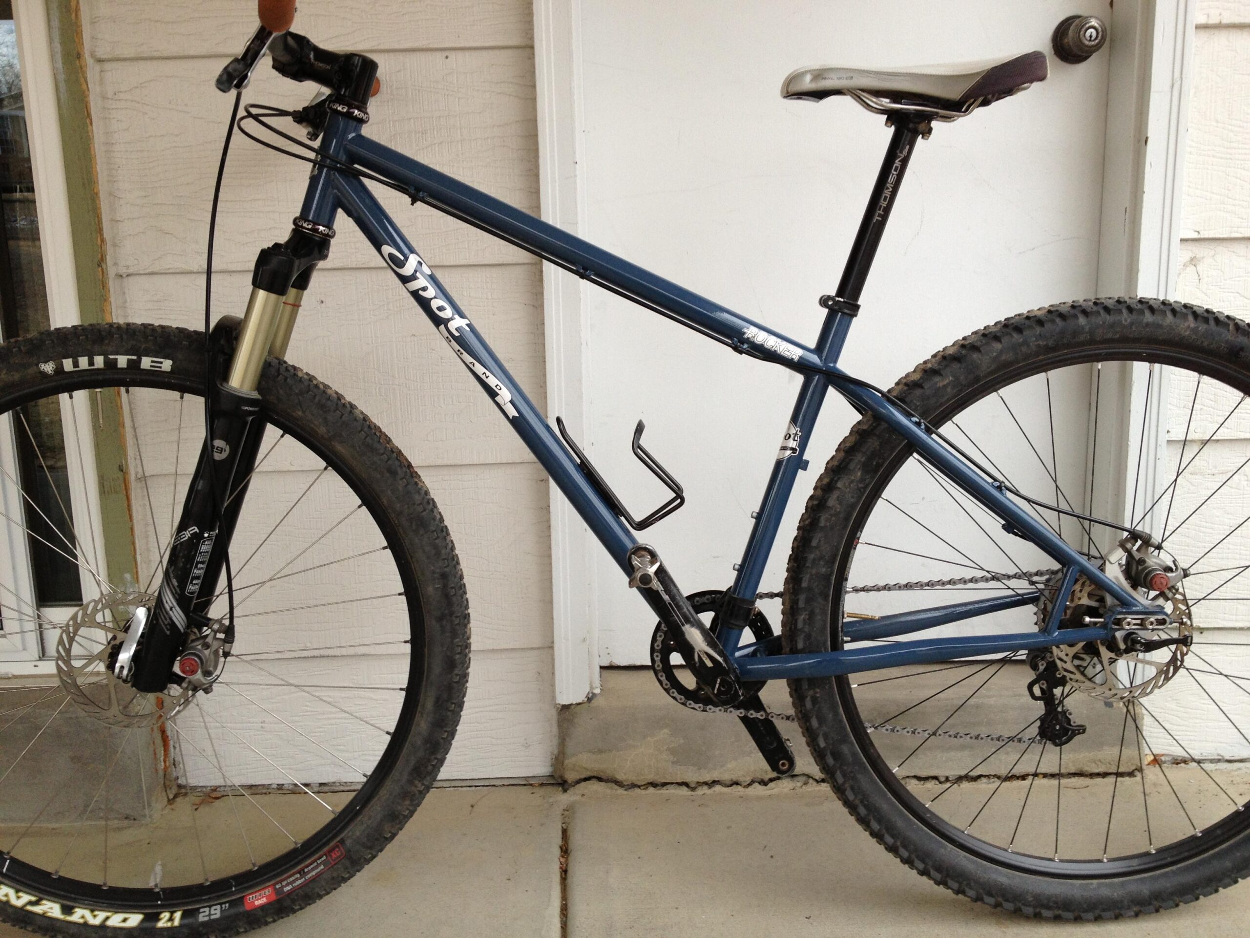 Spot Rocker Spot: A blue mountain bike with a front suspension fork, disc brakes, and knobby tires, resting on a concrete surface in front of a door. The bike features a prominent brand logo on the frame and a sleek design suitable for off-road cycling.