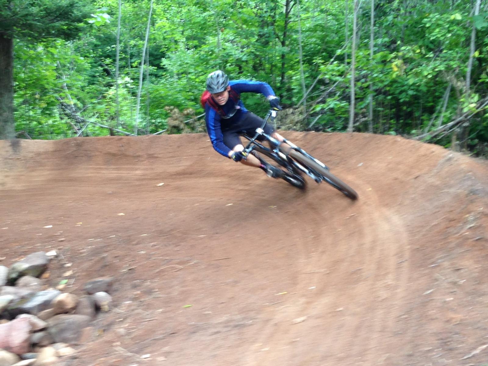 A mountain biker leans into a dirt bank while navigating a turn on a trail surrounded by lush green vegetation. The rider is wearing a helmet and protective gear, with a focused expression, showing skill and determination. Rocks are visible on the side of the path. WinMan Trails mountain bike trail.