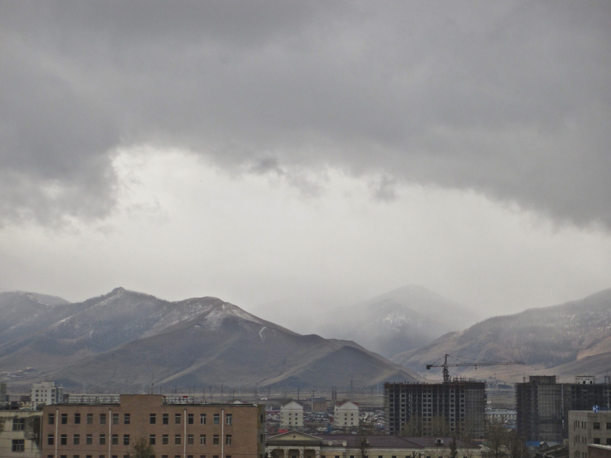 A cloudy sky over a mountainous landscape, featuring snow-capped peaks in the distance. In the foreground, there are several buildings, including a construction site, with various architectural styles visible. The atmosphere appears overcast, suggesting a chilly or damp weather. Zaisan Chutes mountain bike trail.