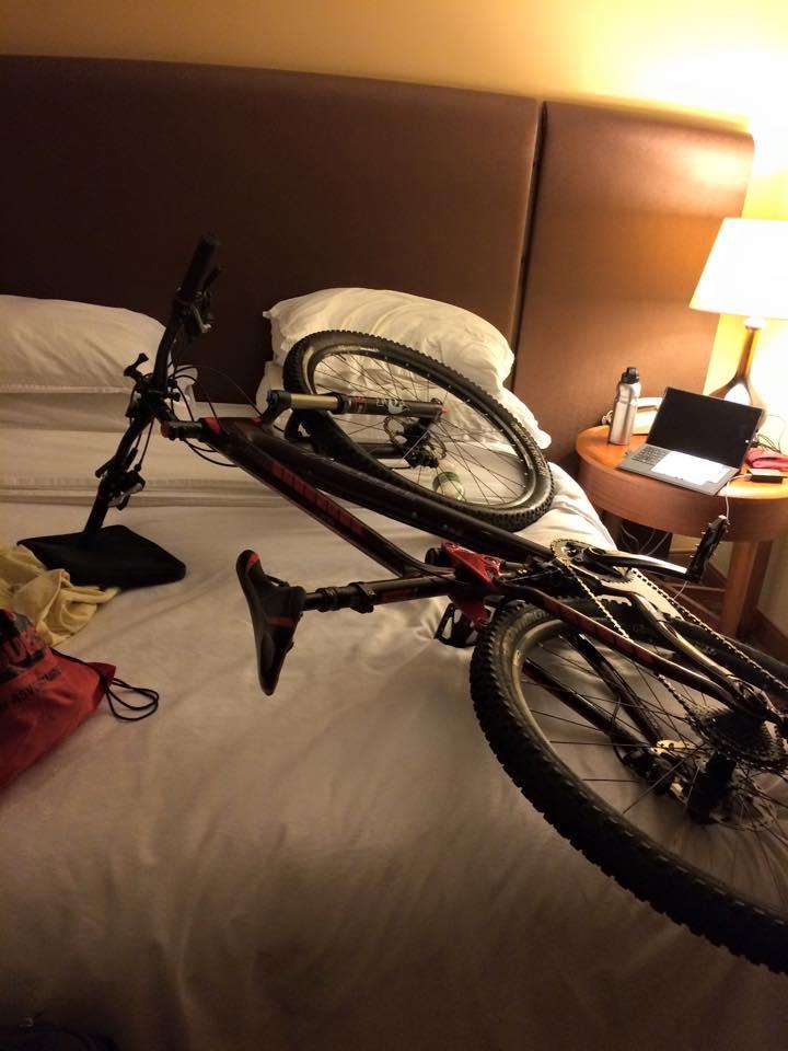 A mountain bike is placed on a bed in a hotel room, with pillows and a headboard in the background. A laptop and water bottle are visible on a side table next to the bed. Alafia River State Park mountain bike trail.