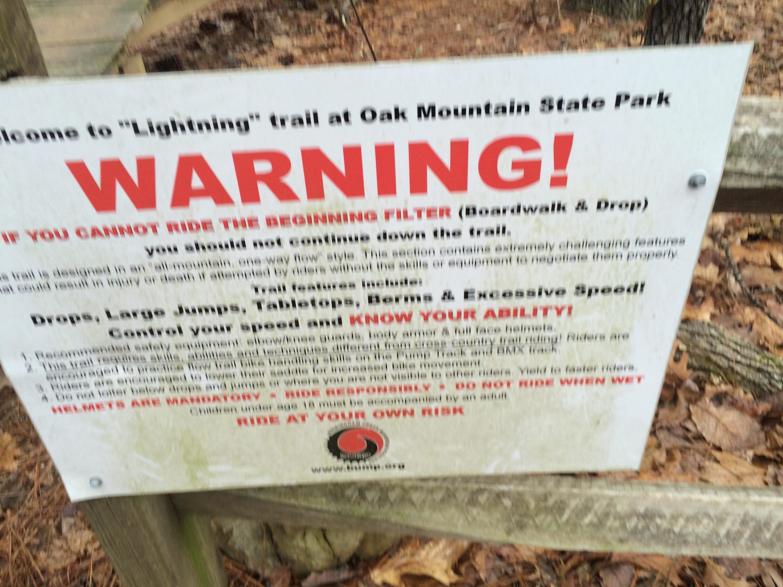 A cautionary sign at Oak Mountain State Park indicating that the "Lightning" trail includes challenging features such as large jumps and speed requirements. The sign warns that riders who cannot navigate the beginning filter should not proceed and emphasizes the importance of wearing safety gear and controlling speed. It also states that helmets are mandatory and recommends caution in wet conditions. The background shows a natural setting with fallen leaves and wooden fencing. Oak Mountain State Park mountain bike trail.