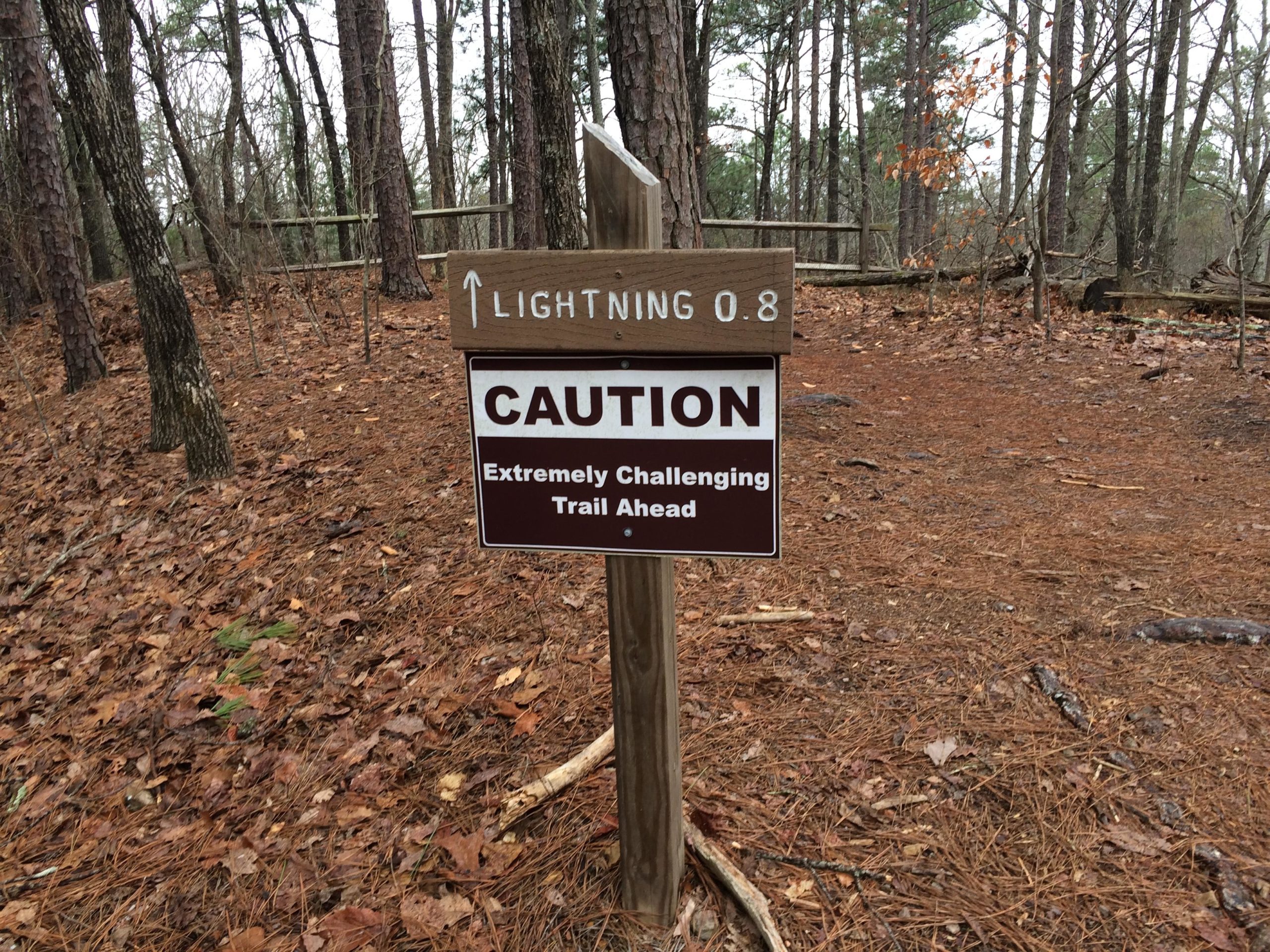 Caution sign indicating an extremely challenging trail ahead, with a directional arrow pointing towards "Lightning 0.8." The trail is surrounded by trees and a forested area covered with pine needles and fallen leaves. Oak Mountain State Park mountain bike trail.