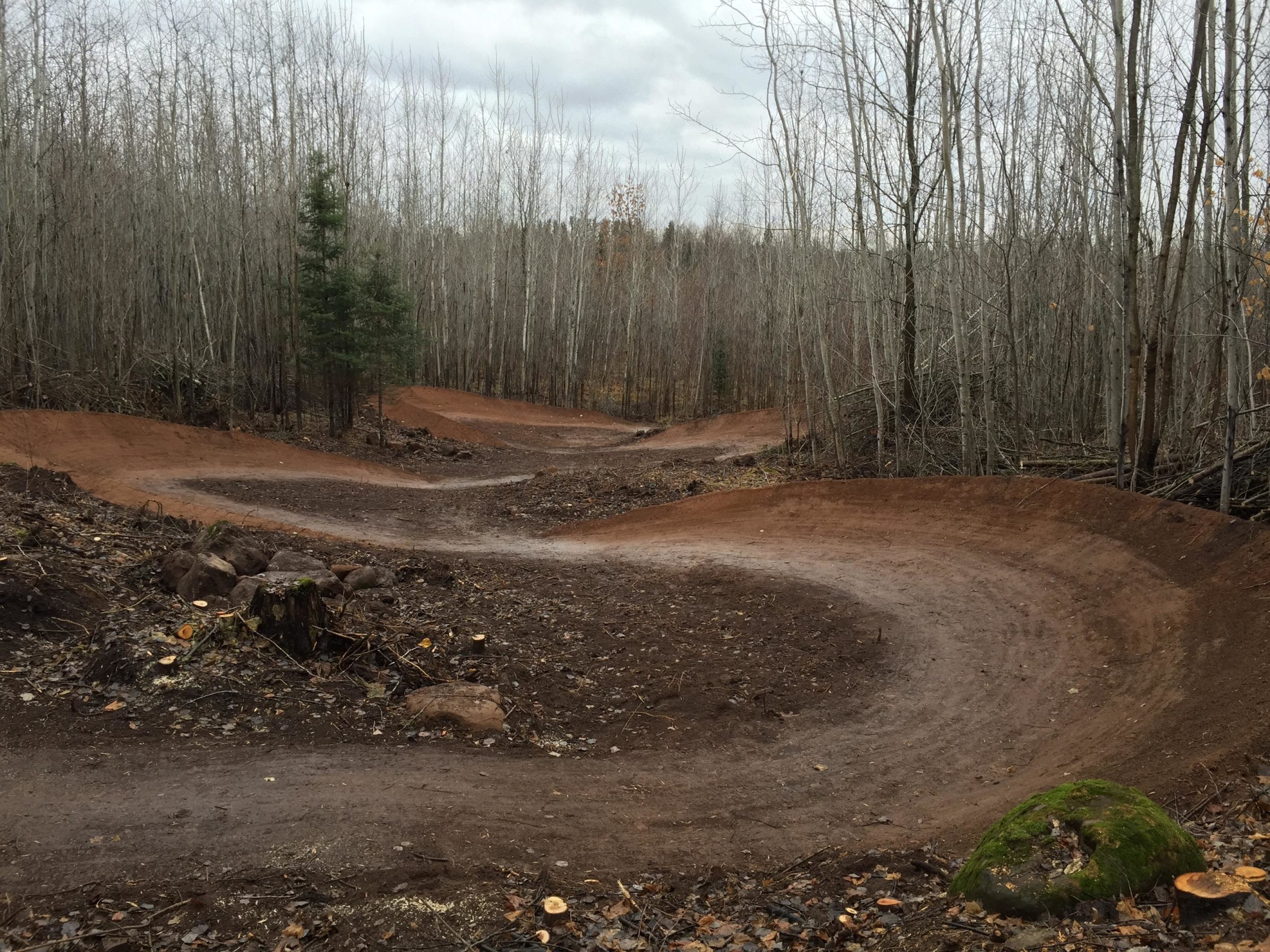 A winding dirt bike trail surrounded by bare trees, with patches of earthy brown soil and scattered rocks. The trail features smooth curves and slight inclines, indicating an area designed for cycling or off-road biking. The cloudy sky above suggests a cool, overcast day. WinMan Trail mountain bike trail.