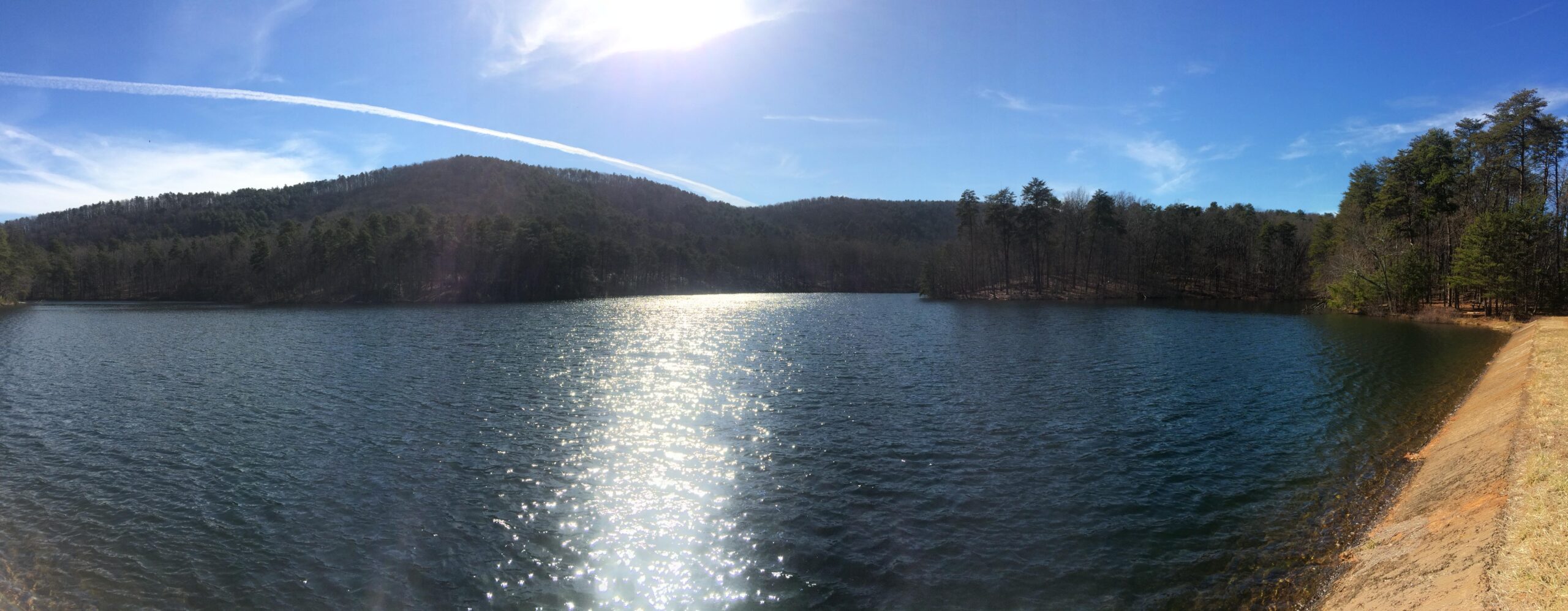 A panoramic view of a serene lake surrounded by rolling hills and lush trees under a clear blue sky. The sun reflects off the water's surface, creating a sparkling effect, while a few wispy clouds trail across the sky. The landscape conveys a sense of calm and natural beauty. Paris Mountain State Park mountain bike trail.