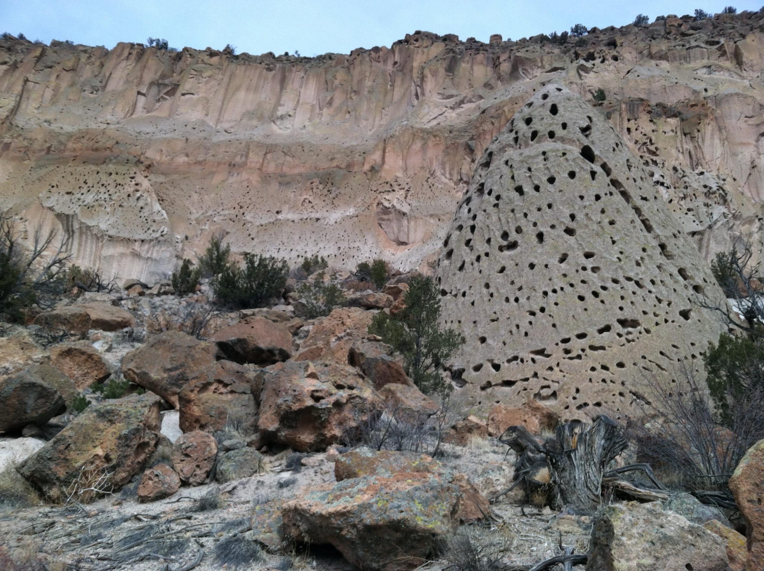 A rocky landscape featuring unique, porous formations. In the foreground, a conical rock structure with numerous holes stands out against a backdrop of layered cliffs and boulders, with sparse vegetation scattered throughout. The sky is overcast, adding a subdued atmosphere to the scene. Pueblo Canyon mountain bike trail.