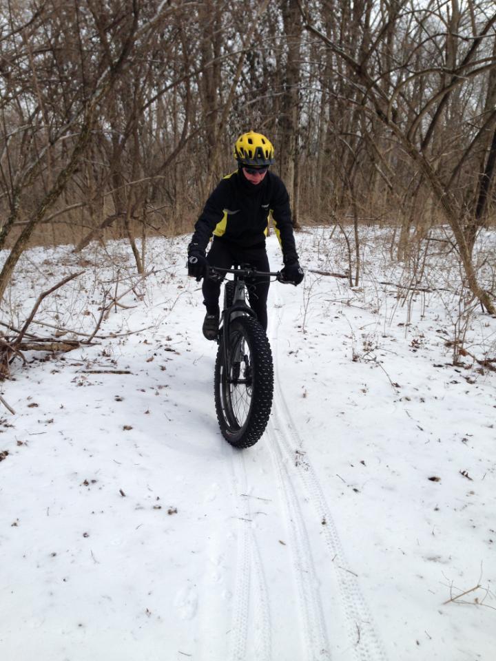 A person wearing a yellow helmet and black cycling gear rides a fat bike on a snowy trail surrounded by bare trees. The ground is covered in snow, and bike tire tracks are visible in the snow. Percy Warner Mountain Bike Trails mountain bike trail.