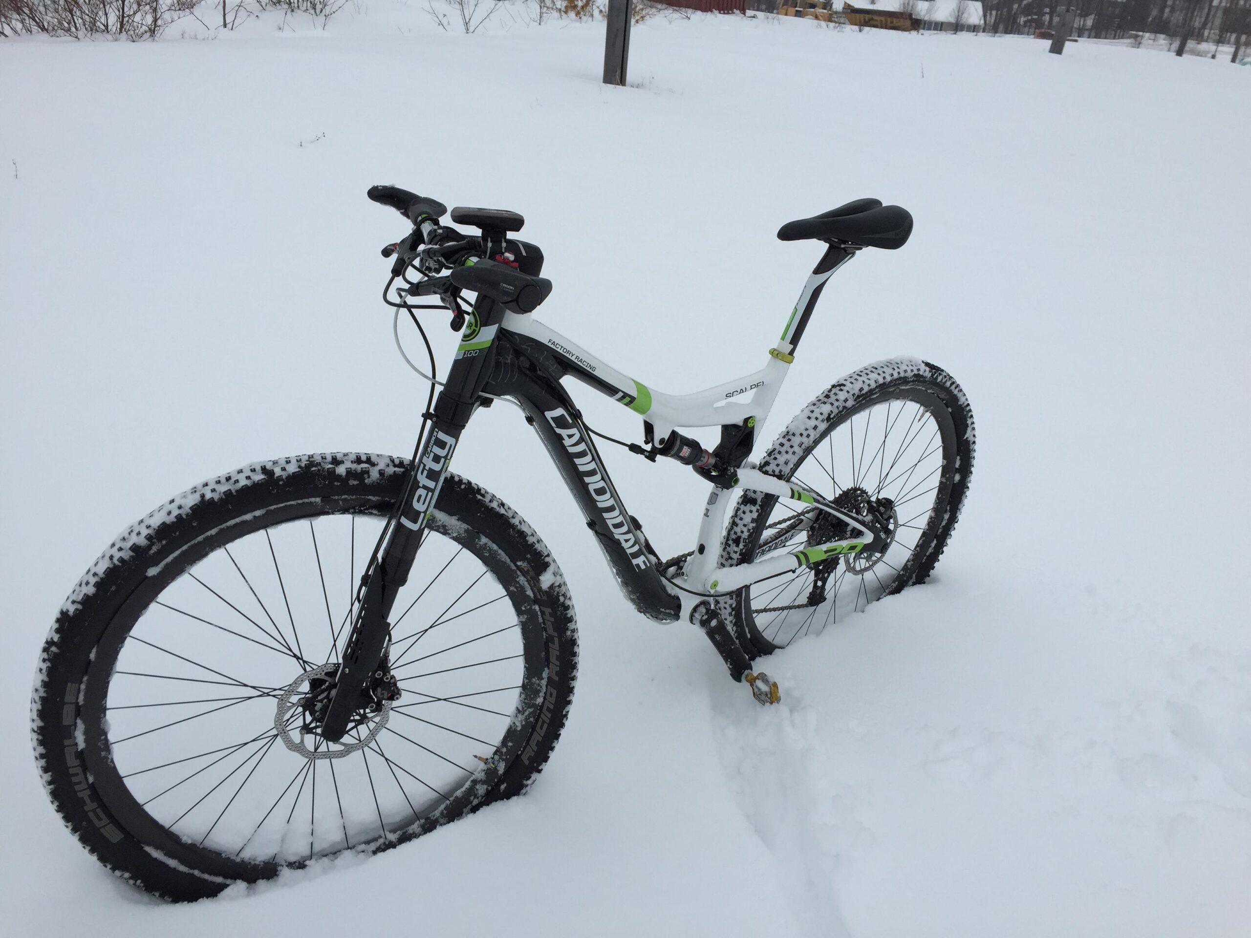 A mountain bike with wide tires is resting on a thick layer of snow. The bike features a black and white frame with green accents, and the tires are partially covered in snow. The background is a snowy landscape with minimal visibility of trees and structures.