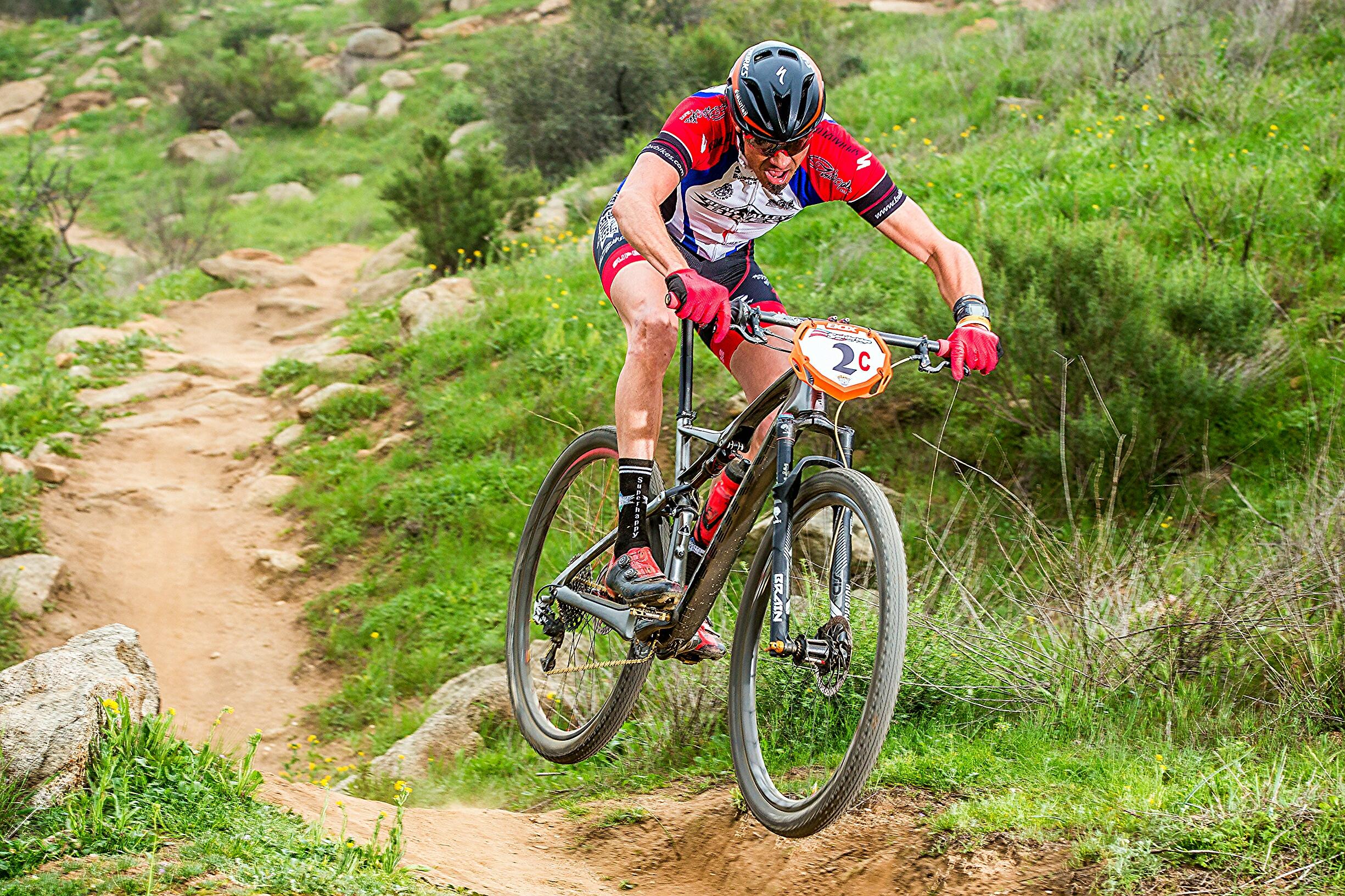 Specialized S-Works Epic WC: A mountain biker in a red and blue jersey performing a jump on a dirt trail surrounded by green grass and rocky terrain. The cyclist is focused and wears a helmet and gloves, with a race number displayed on the bike.