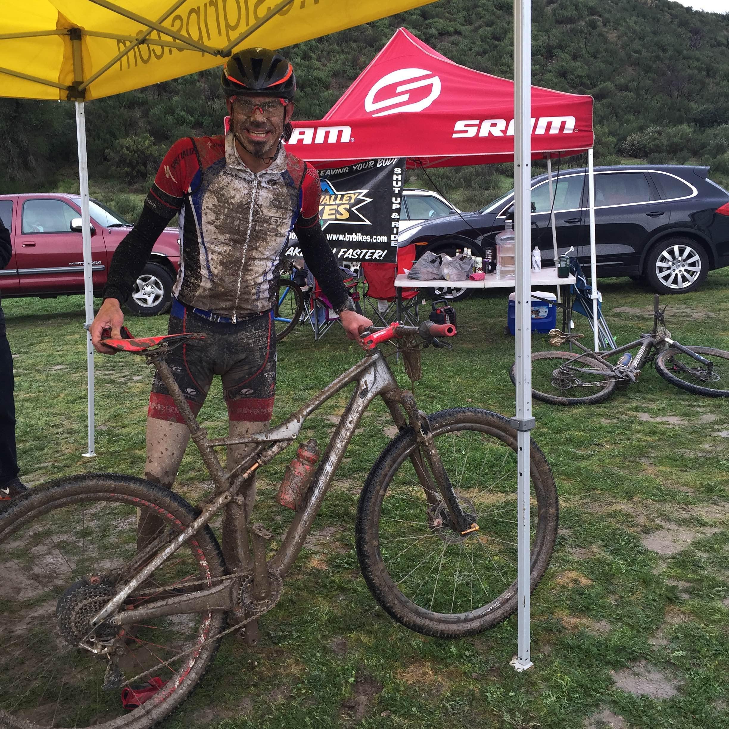 Specialized S-Works Epic WC: A muddy mountain biker stands next to a dirt-covered bike under a tent, smiling after a race. The cyclist is wearing a wet, soiled jersey and shorts, while the bike showcases splatters of mud. In the background, there are vehicles and supplies associated with the event, highlighting a recreational outdoor setting.