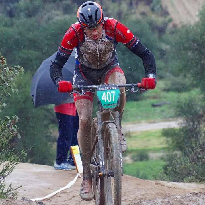 Specialized S-Works Epic WC: A mountain biker covered in mud accelerates through a wet race course, showcasing determination and skill. In the background, a person holds an umbrella, emphasizing the rainy conditions. The biker's number, 407, is clearly visible on their racing jersey.