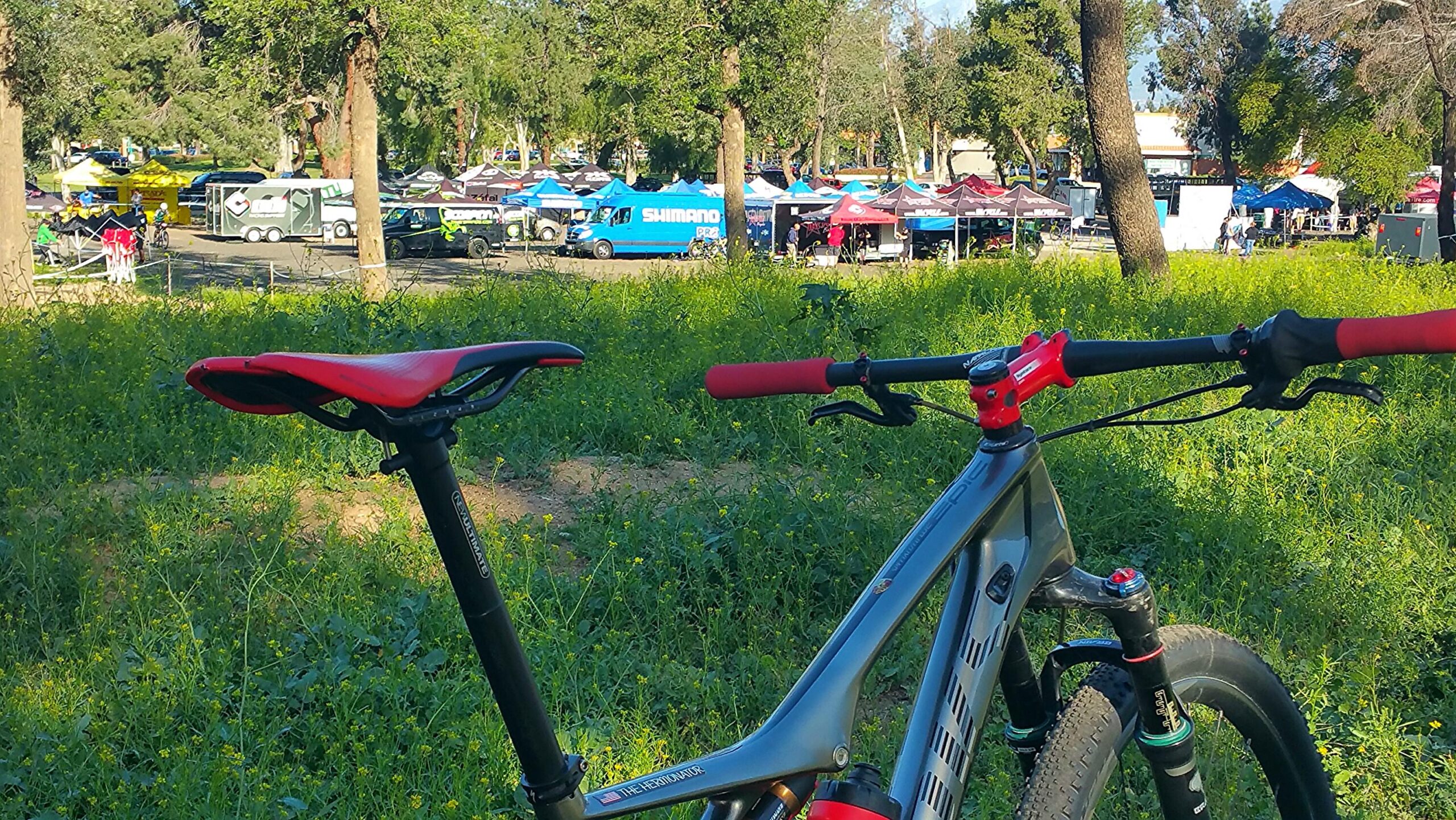 Specialized S-Works Epic WC: A close-up view of a mountain bike with a red saddle and handlebars, positioned in a grassy area. In the background, various colorful tents and vehicles are set up for an outdoor event, surrounded by trees.