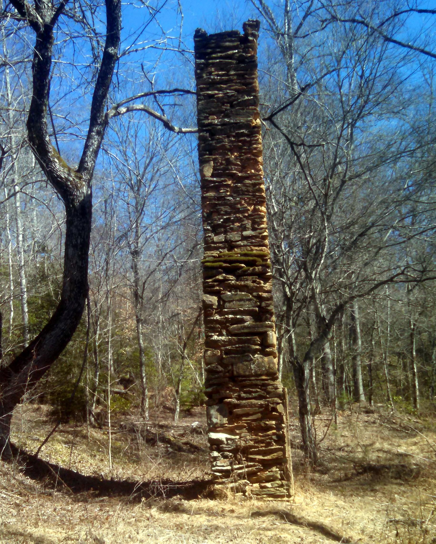 A tall, weathered stone chimney stands alone in a clearing surrounded by bare trees and dry grass, under a clear blue sky. Tsali Left Loop mountain bike trail.