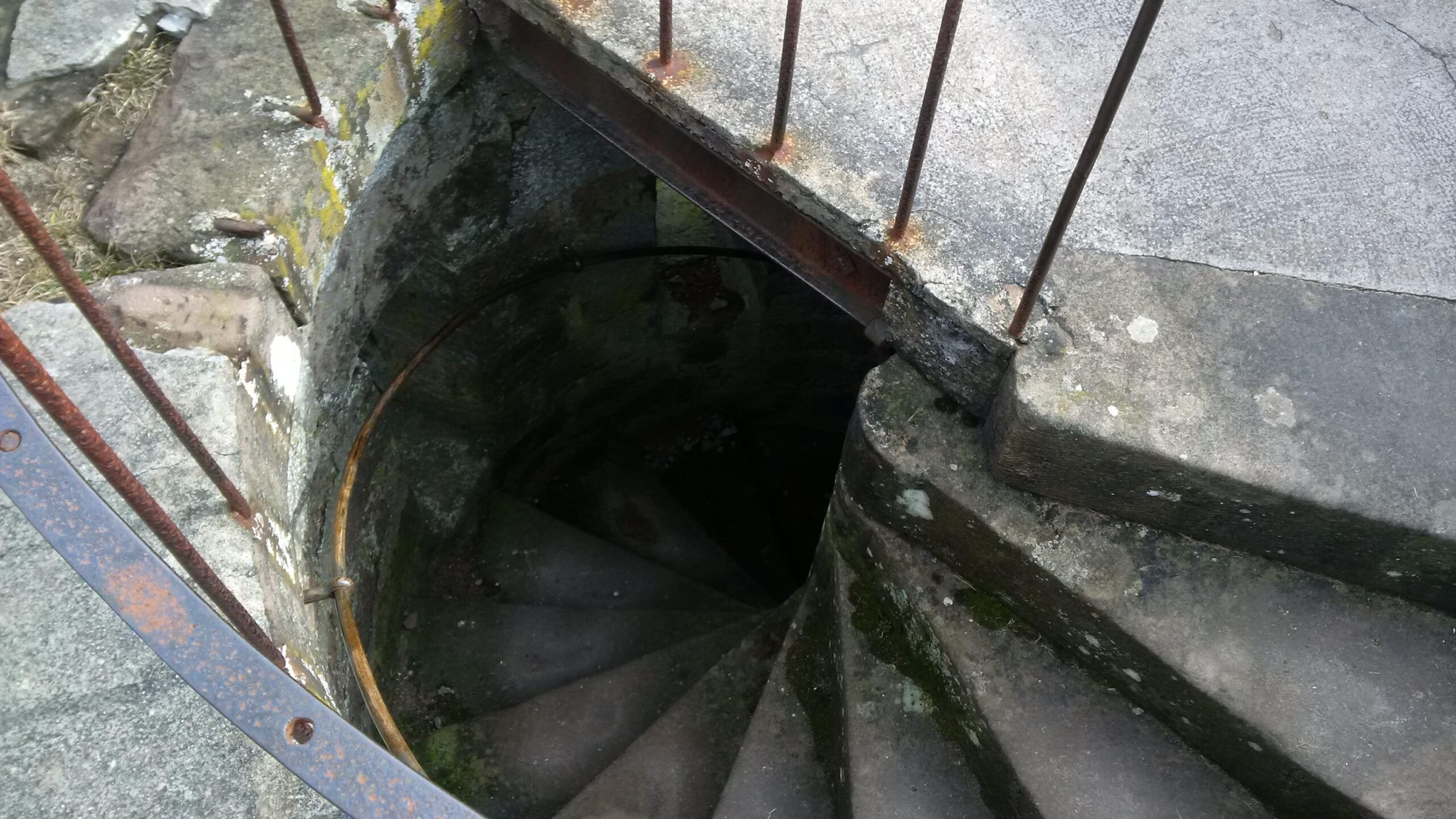 Alt text: A view looking down a spiral stone staircase with moss on the steps, bordered by rusty metal railings, leading into a darkened well or underground space. Johanniskreuz Trail 9 mountain bike trail.