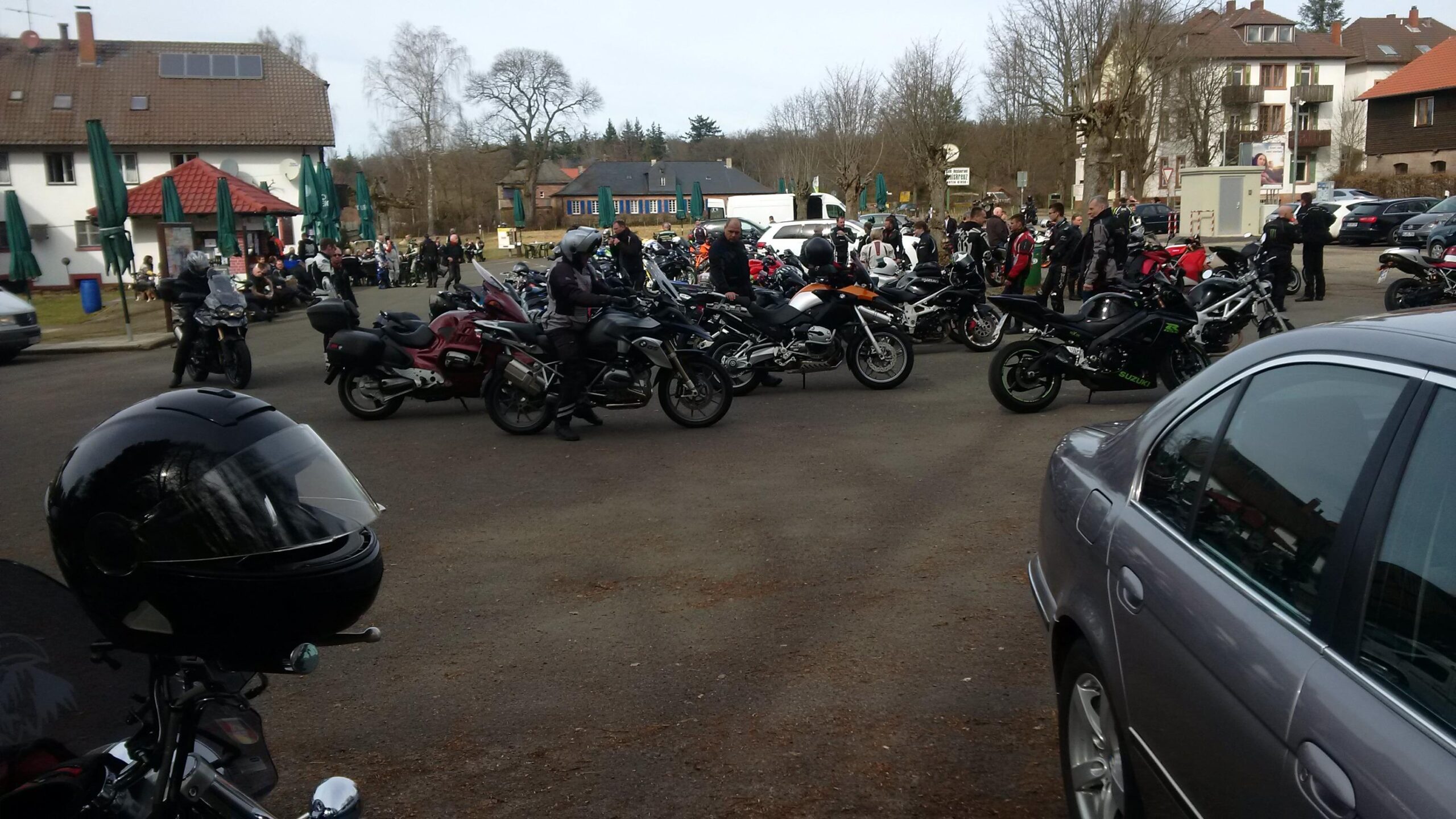 A busy parking lot filled with various motorcycles and riders, with some people gathered around benches outside a building. Trees and cars are visible in the background, creating a lively outdoor scene. Johanniskreuz Trail 9 mountain bike trail.