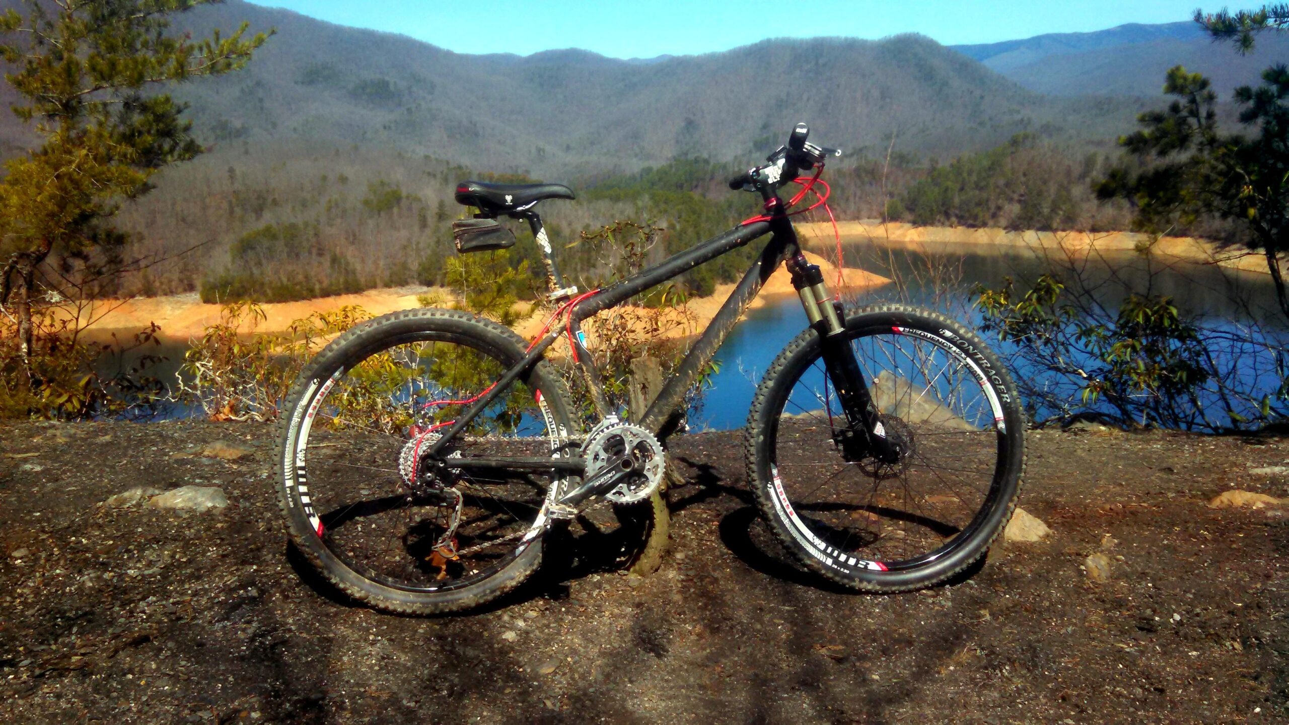 A mountain bike parked on a rocky overlook with a scenic view of a lake and mountains in the background. The bike has a black frame with red accents and is positioned on a dirt path surrounded by sparse vegetation. The calm water of the lake reflects the clear blue sky and the distant hills. Tsali Left Loop mountain bike trail.
