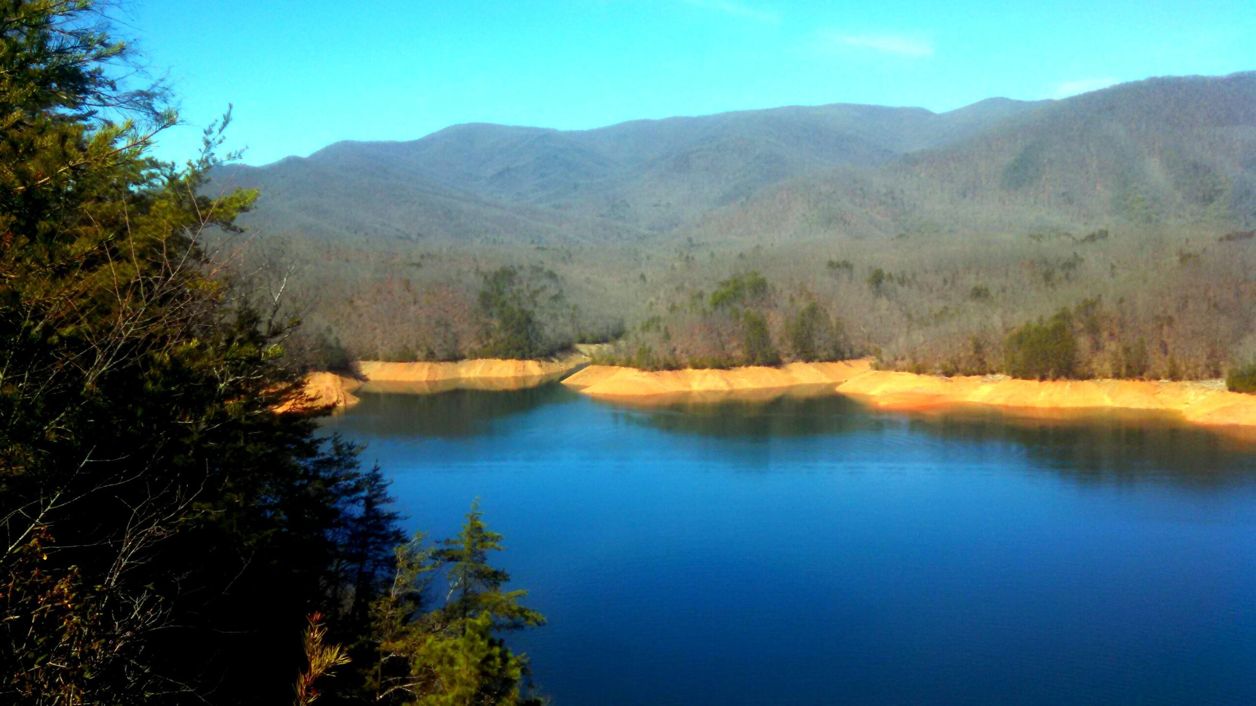 A serene landscape featuring a calm lake surrounded by verdant trees and rolling mountains. The water reflects the blue sky, while the distant hills are adorned with a mix of bare and green trees, emphasizing the peacefulness of the natural scenery. Tsali Left Loop mountain bike trail.