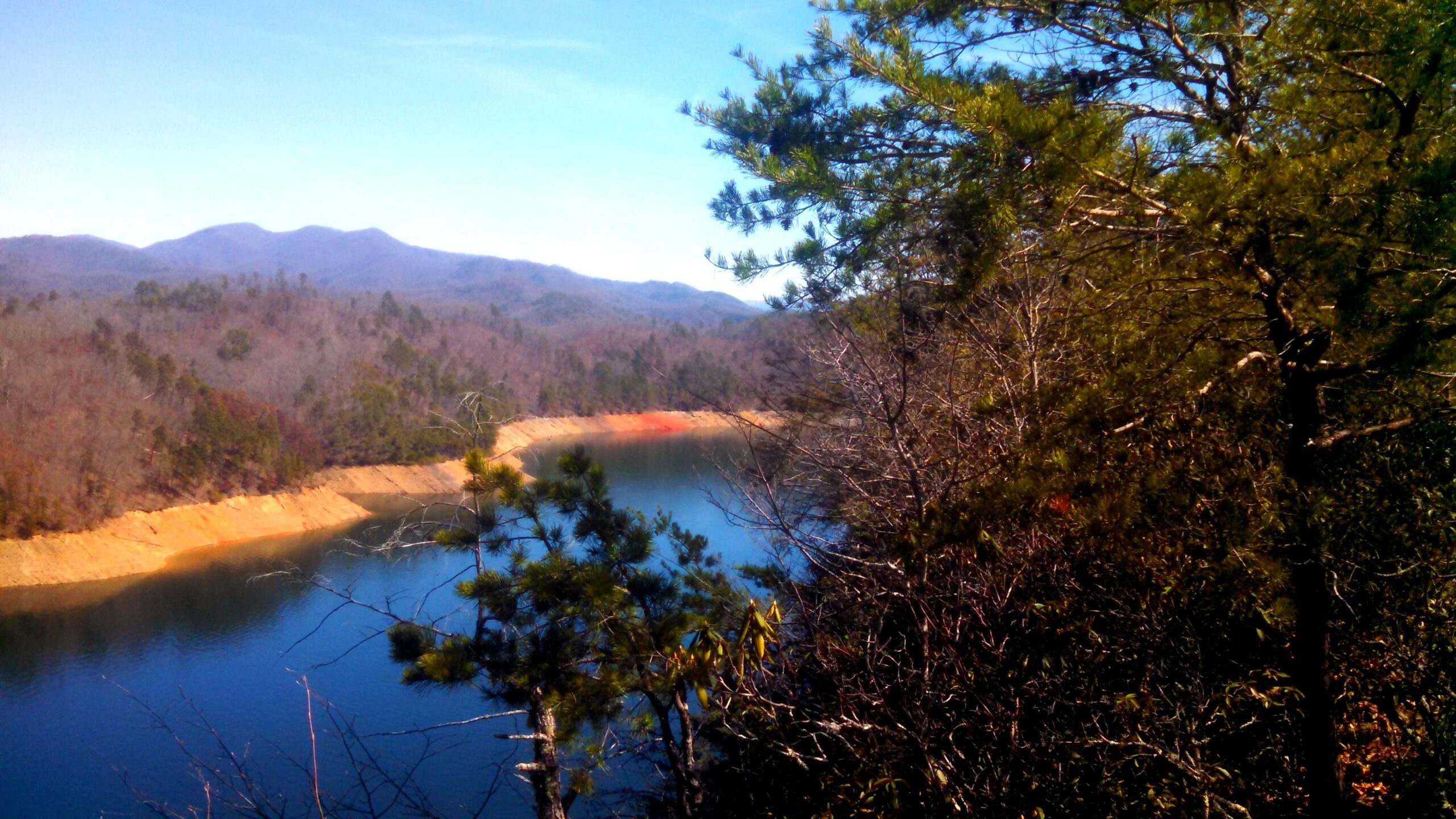 A serene landscape showing a calm lake surrounded by mountains, with trees and shrubs in the foreground. The sky is clear and blue, reflecting off the water's surface. The shoreline features a mix of sandy and earthy colors, adding to the natural beauty of the scene. Tsali Left Loop mountain bike trail.