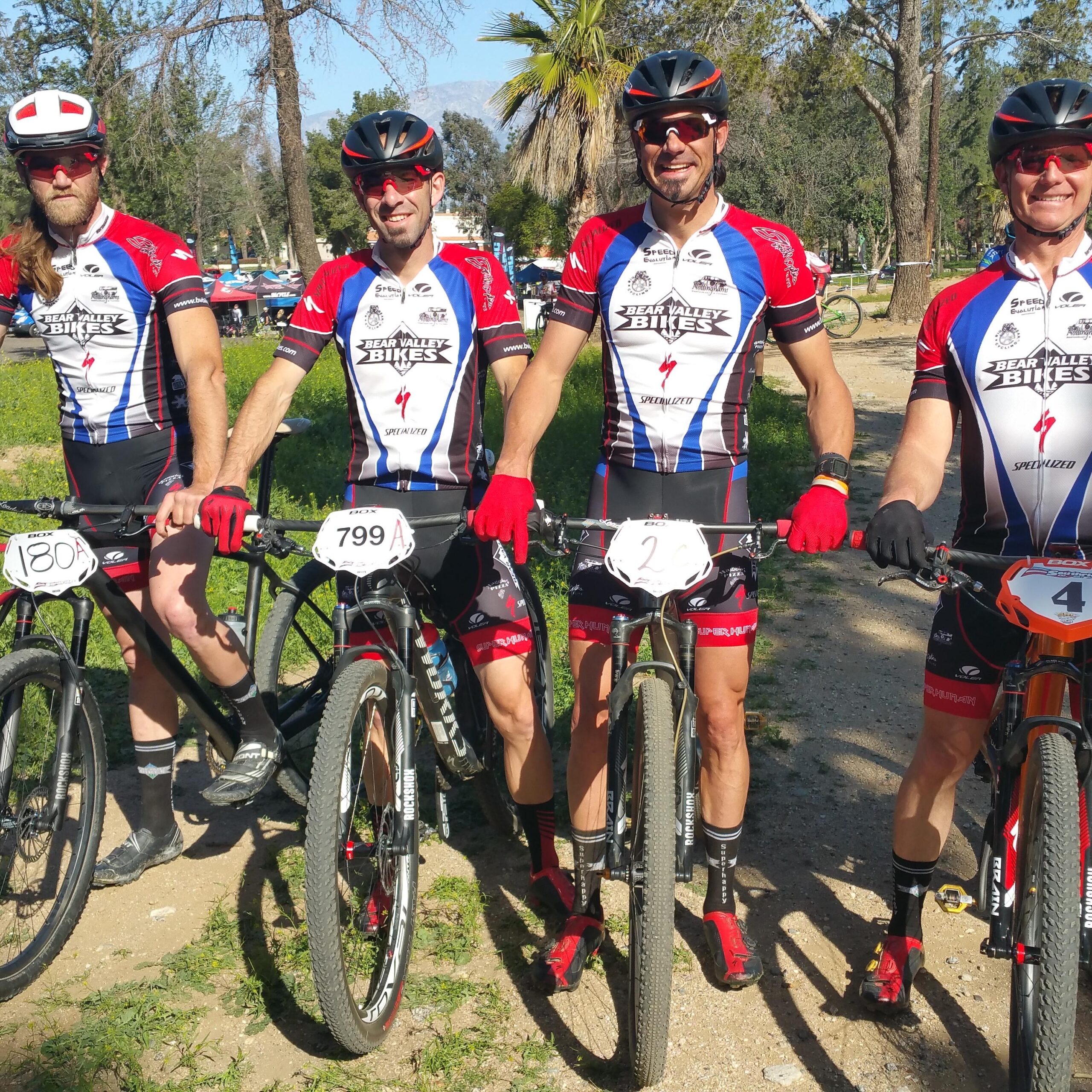 Specialized S-Works Epic WC: Four male mountain bikers pose for a photo in a sunny outdoor setting. They are wearing matching cycling jerseys featuring red, white, and blue colors with the logo "Bear Valley Bikes." Each cyclist is holding a bike and displaying their race numbers: 180, 799A, and 2. The background shows greenery and trees, indicating a race or event location.