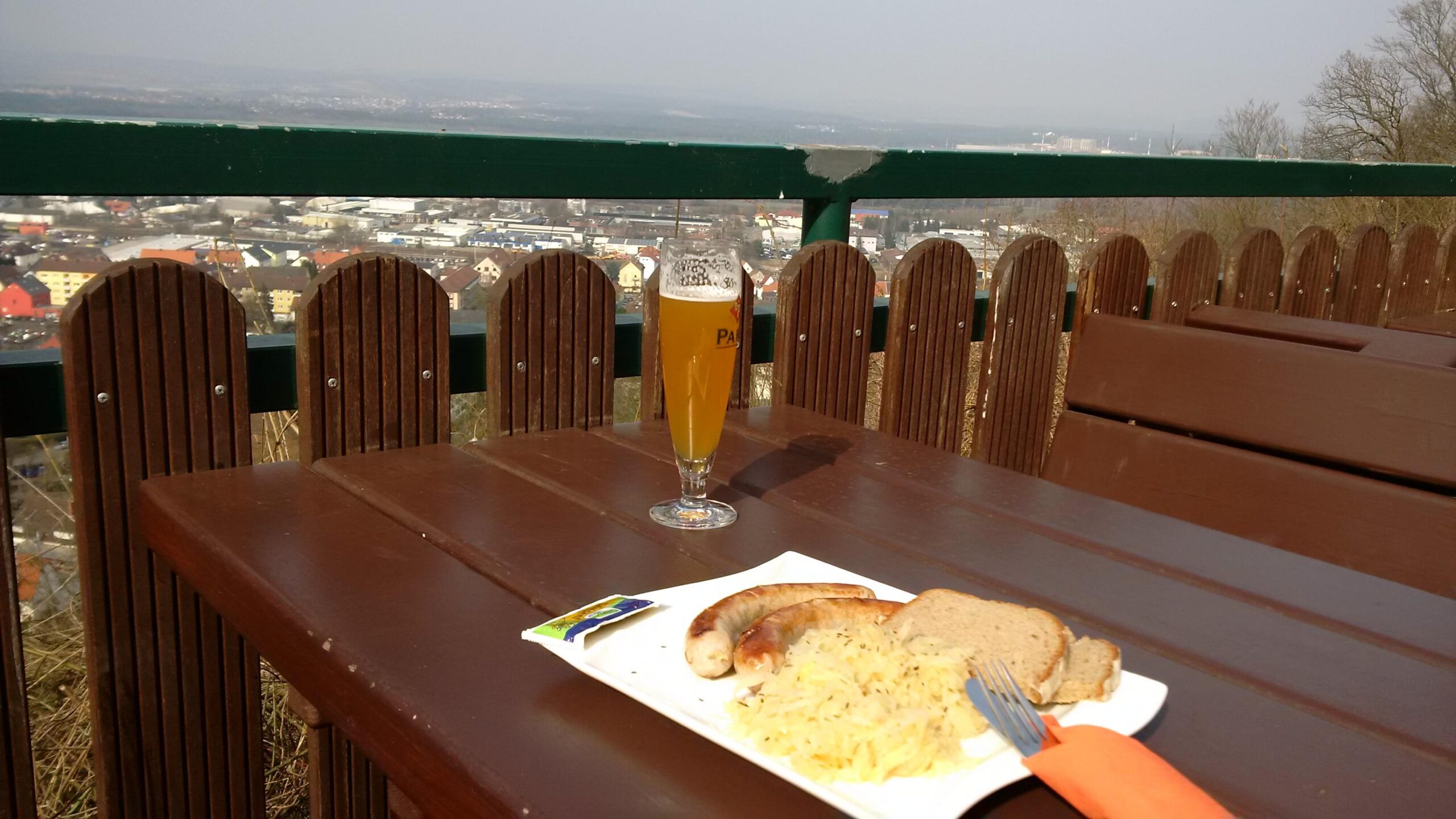 A table setting featuring a glass of beer and a plate of food, including sausages, sauerkraut, and bread, with a scenic view of a town in the background under a clear sky. Landstuhl Trail mountain bike trail.