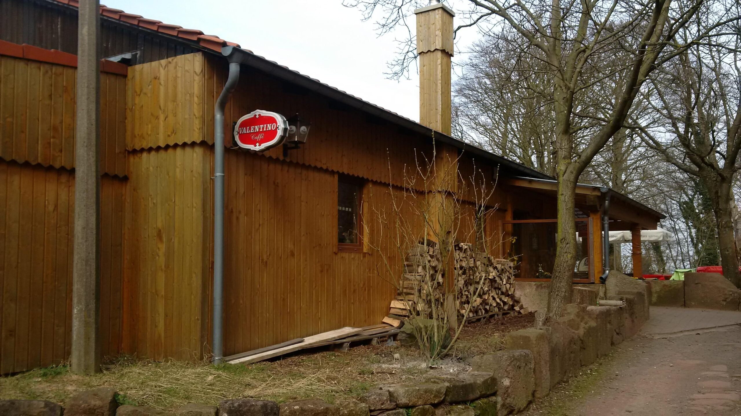 A wooden building with a sloped roof, featuring a sign that reads "Valentino Caffè." The exterior has natural wood paneling and a chimney, with firewood stacked beside it. Surrounding the building are bare trees and a pathway made of stones. The scene conveys a rustic, inviting atmosphere. Landstuhl Trail mountain bike trail.