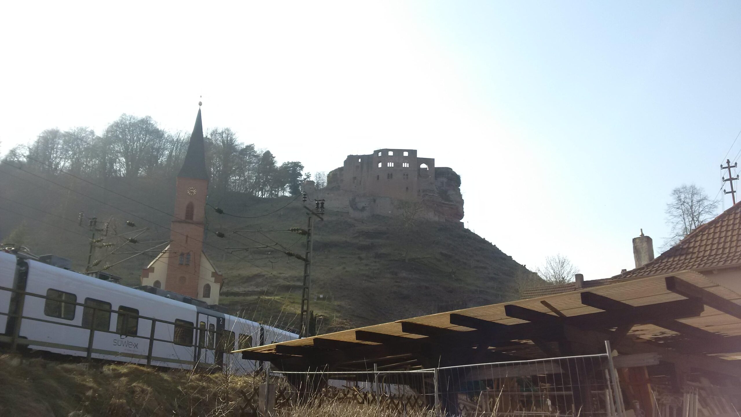 A scenic view featuring a church with a tall spire in the foreground, a passing train, and a historic castle ruin perched on a hill in the background. The landscape is bathed in soft sunlight, with trees lining the hillsides. Ortsgemeinde Frankenstein mountain bike trail.