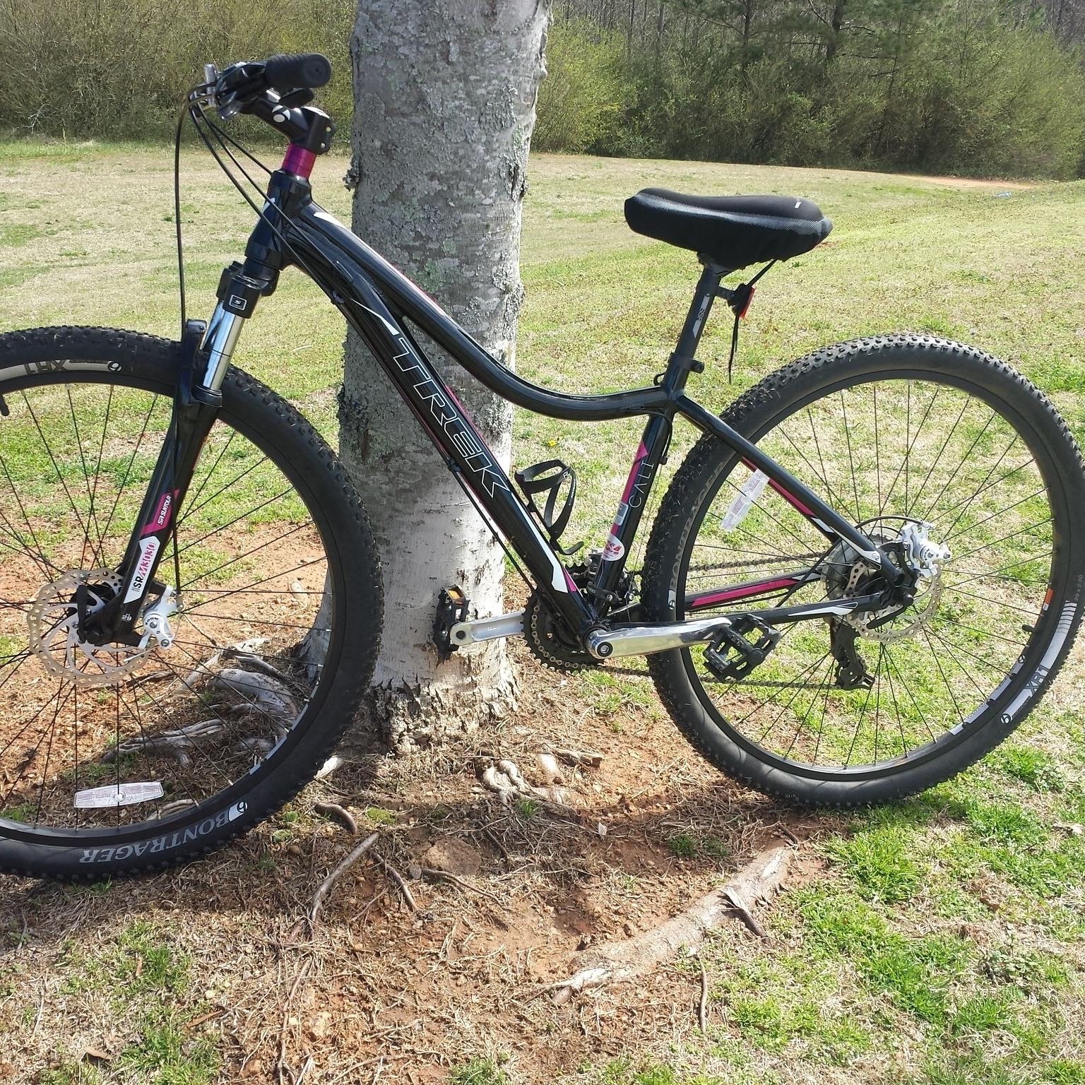 Trek Cali: A black and pink mountain bike leaning against a tree, set in a grassy area with sparse trees in the background. The bike features knobby tires and a comfortable saddle, indicating it's designed for off-road riding.