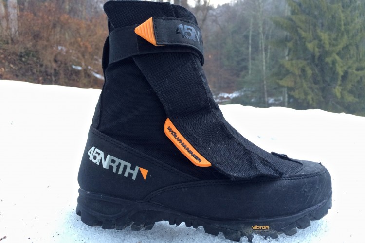 A close-up image of a black snow boot with a rugged sole, featuring an adjustable strap and orange accents. The boot is designed for winter conditions, set against a snowy outdoor backdrop. The brand "45NRTH" is visible on the side of the boot.