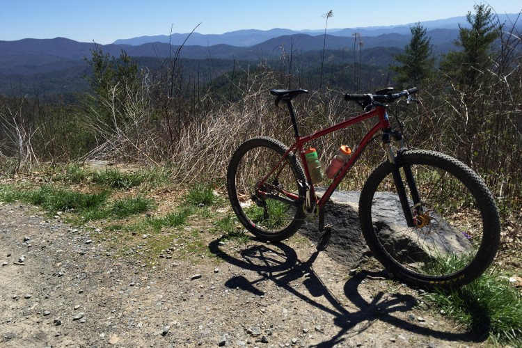 A red mountain bike parked on a rocky outcrop overlooking a scenic view of rolling mountains and a clear blue sky, surrounded by green grass and sparse vegetation.