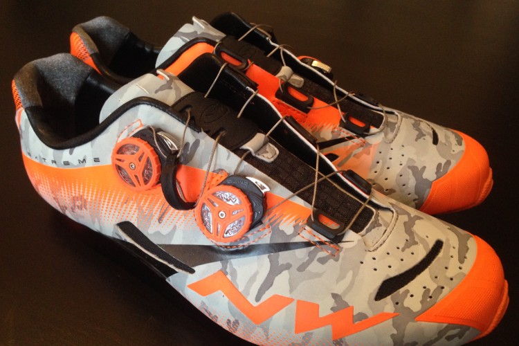 A pair of cycling shoes featuring a grey and orange camouflage design, with a sleek, aerodynamic shape. The shoes have two prominent closure dials for a secure fit and a textured sole optimized for pedal grip. They are presented on a dark surface, showcasing their modern style and athletic functionality.