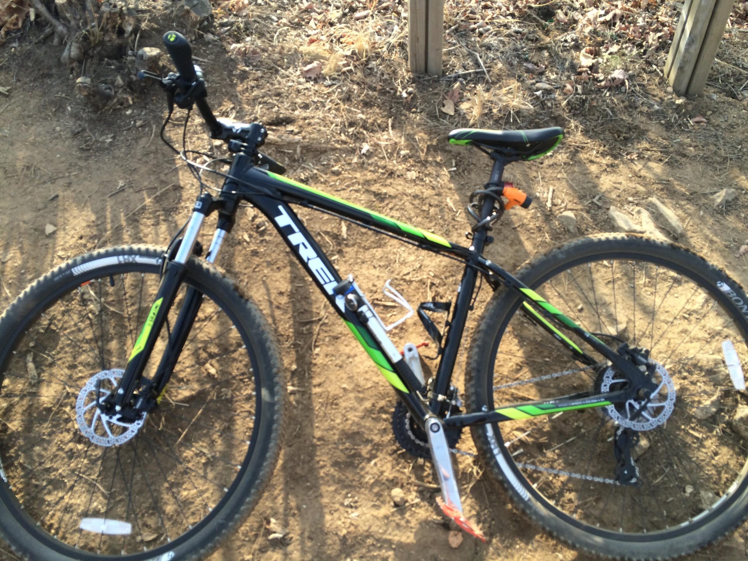 Trek Marlin 6: A black and green mountain bike resting on dirt terrain, featuring disc brakes and a sturdy frame. The bike is positioned at an angle, showing the front wheel and handlebars. In the background, there are patches of soil and scattered leaves.