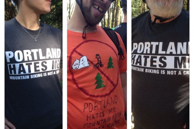 Three individuals are shown wearing T-shirts with a humorous theme related to Portland and mountain biking. The person on the left wears a gray shirt that reads "PORTLAND HATES ME" with "MOUNTAIN BIKING IS NOT A CRIME" underneath. The person in the center has an orange shirt featuring a playful drawing of a mountain biker, along with a similar slogan. The individual on the right also sports a gray shirt with the same text as the first person's shirt. All participants are outdoors with a forest background.