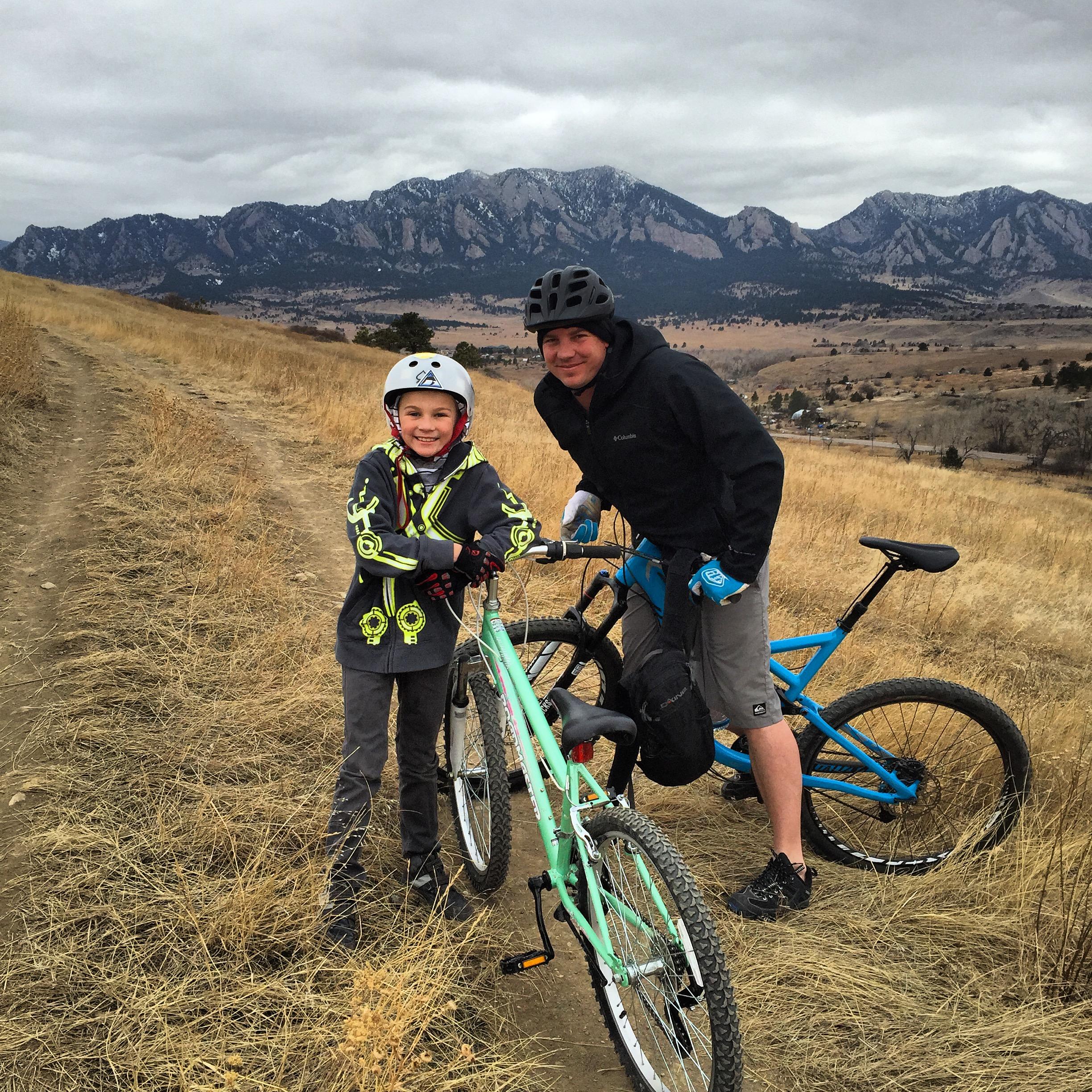 A child and an adult stand beside their bicycles on a dirt path in a grassy area, with mountains in the background under a cloudy sky. The child wears a white helmet and a gray sweatshirt with bright designs, while the adult is dressed in a black jacket and shorts. Both are smiling and appear to be enjoying a day of biking outdoors. Marshall Road Open Spaces mountain bike trail.