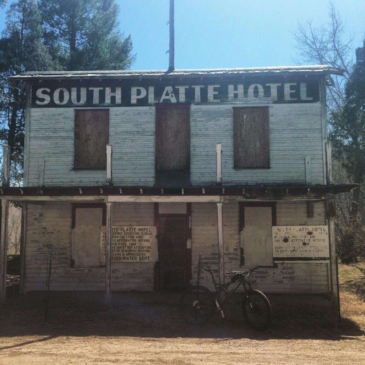 An old, weathered building with a sign reading "South Platte Hotel." The structure has boarded-up windows, peeling paint, and various notices on its exterior. A black bicycle leans against the front of the building, and the scene is set under a clear blue sky with trees in the background. North Fork mountain bike trail.