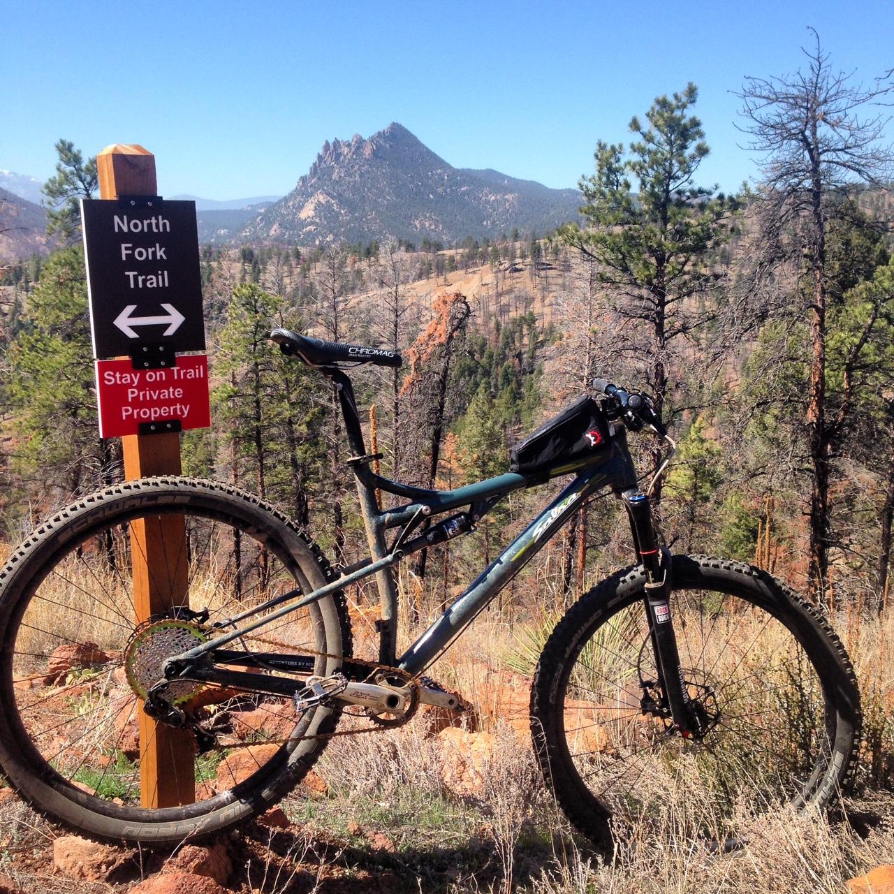 Salsa Horsethief: A mountain bike is leaning against a wooden sign for the North Fork Trail, which has arrows indicating direction and a note to stay on the trail due to private property. The background features a scenic view of mountains and pine trees under a clear blue sky.