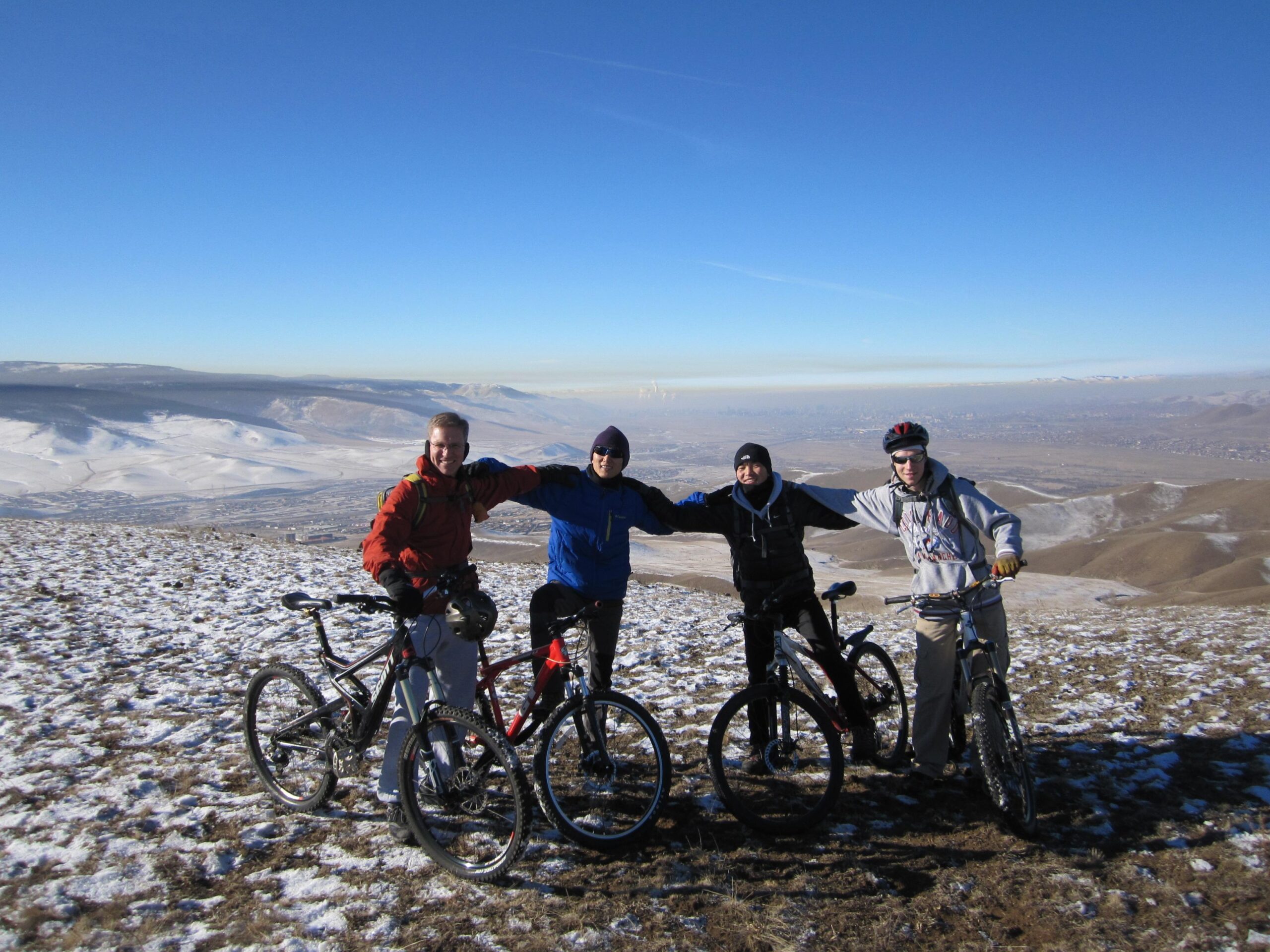 Four mountain bikers are standing on a snowy hillside, each with their bike. They are smiling and posing with their arms around each other, set against a clear blue sky and distant mountains. The landscape features a mix of snow and dry terrain, showcasing a scenic view. Bayanzurkh Uul West mountain bike trail.