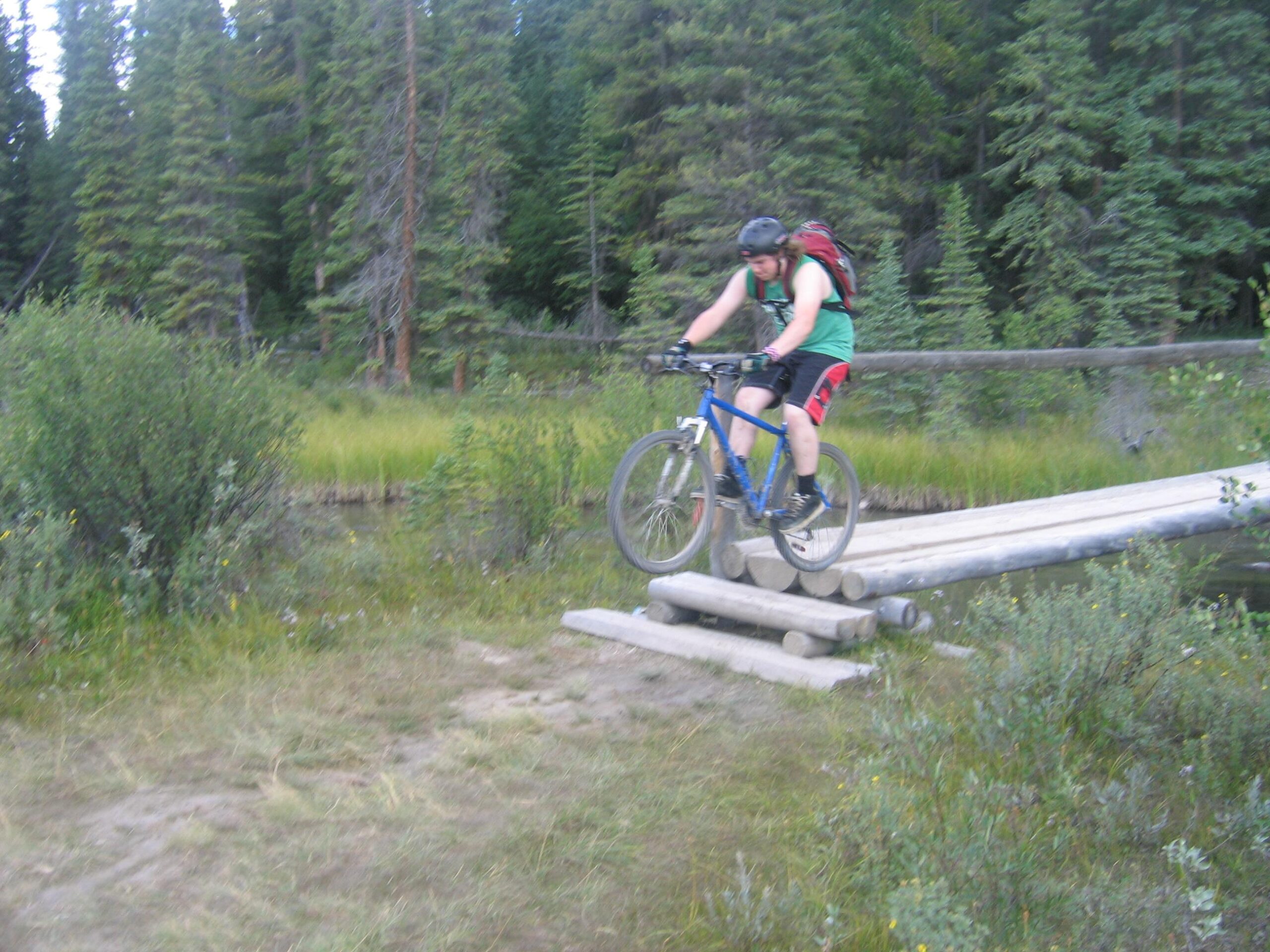 A person in a green tank top and black shorts is riding a bike over a wooden bridge in a forested area. The cyclist is airborne, with one wheel lifted off the ground, surrounded by green grass and trees. The Overlander mountain bike trail.